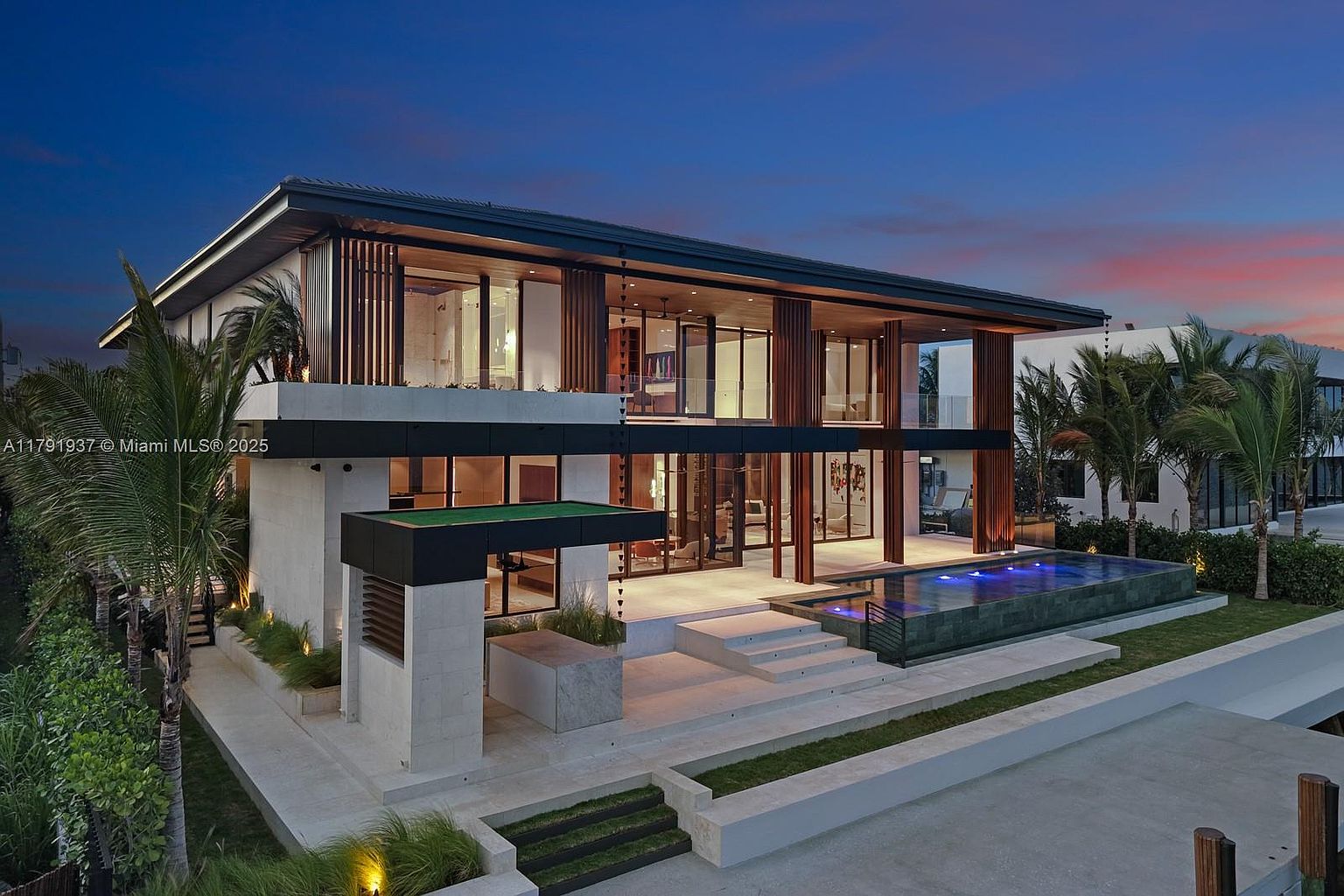 This is a stunning rear view of a modern luxury home at dusk, featuring expansive glass walls, wooden accents, and a sleek, dark-colored roof. A rectangular pool with illuminated water adds a touch of elegance, while the multi-level patio and manicured landscaping create a sophisticated outdoor living space. The architecture emphasizes clean lines and a seamless integration of indoor and outdoor areas.