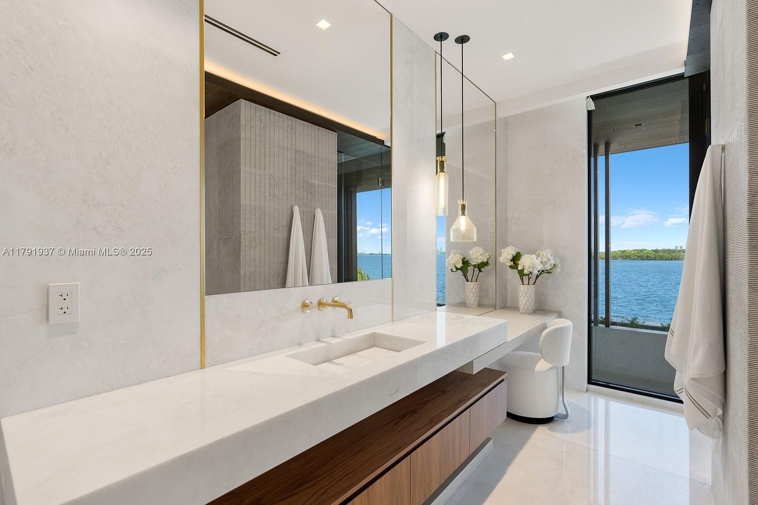 This is a luxurious primary bathroom featuring a long, white marble countertop with dual sinks and gold fixtures. A large mirror with gold trim reflects the space, and pendant lights hang elegantly above the counter. A large window offers a stunning view of the water, and a modern chair sits beneath the counter, creating a spa-like atmosphere.