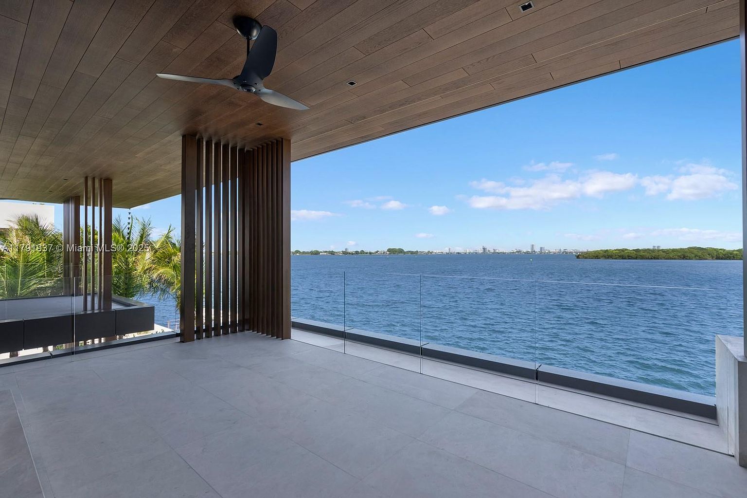 This image showcases a luxurious outdoor balcony with a stunning waterfront view. The space features a sleek, modern design with a wood-paneled ceiling, a contemporary ceiling fan, and a glass railing that provides an unobstructed view of the water. The neutral-toned tile flooring complements the overall aesthetic, creating a serene and sophisticated outdoor living area.