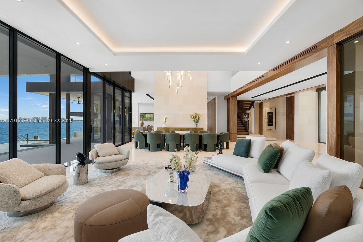 This is a luxurious living room featuring a spacious layout with modern design elements. The room includes a large white sectional sofa with decorative pillows, comfortable armchairs, and a unique coffee table on a plush rug. Floor-to-ceiling windows offer stunning water views, enhancing the room's appeal and creating a bright and airy atmosphere.