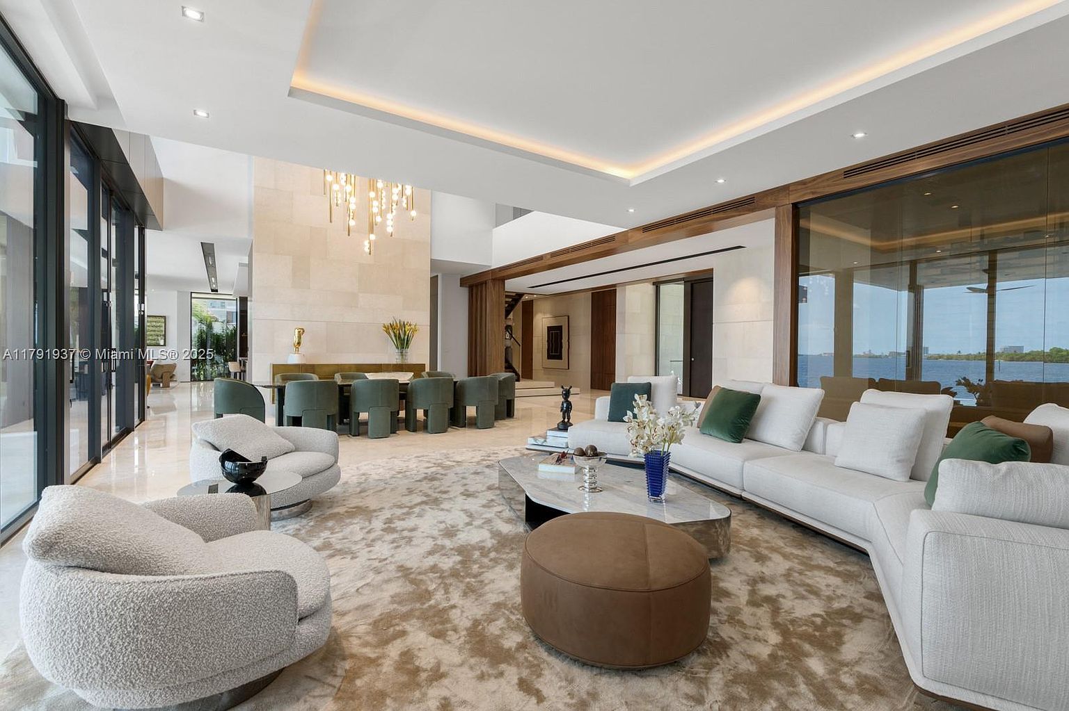 This is a spacious and luxurious living room featuring a neutral color palette with pops of green and blue. The room includes plush seating arrangements, a large area rug, and floor-to-ceiling windows that offer stunning views. A dining area is visible in the background, creating an open and airy atmosphere.