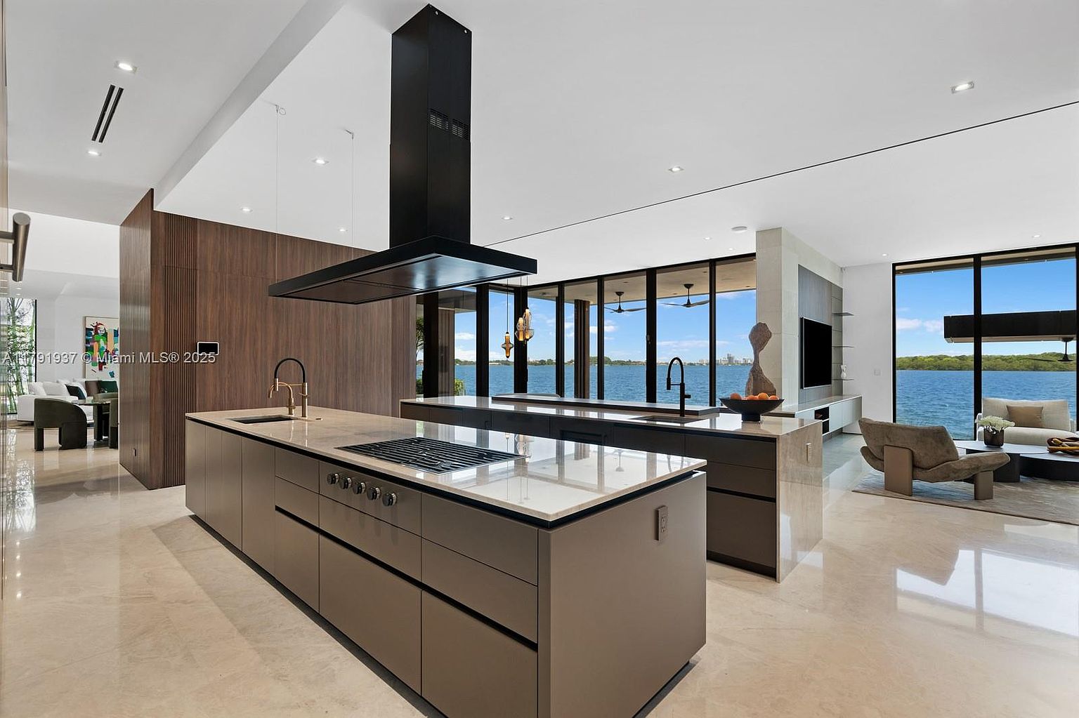 This is a modern, luxurious kitchen featuring two large islands with light countertops and dark cabinetry. A sleek, black range hood hangs above the main island, and floor-to-ceiling windows offer stunning water views. The open-concept design seamlessly connects the kitchen to the living area, creating a spacious and inviting atmosphere.