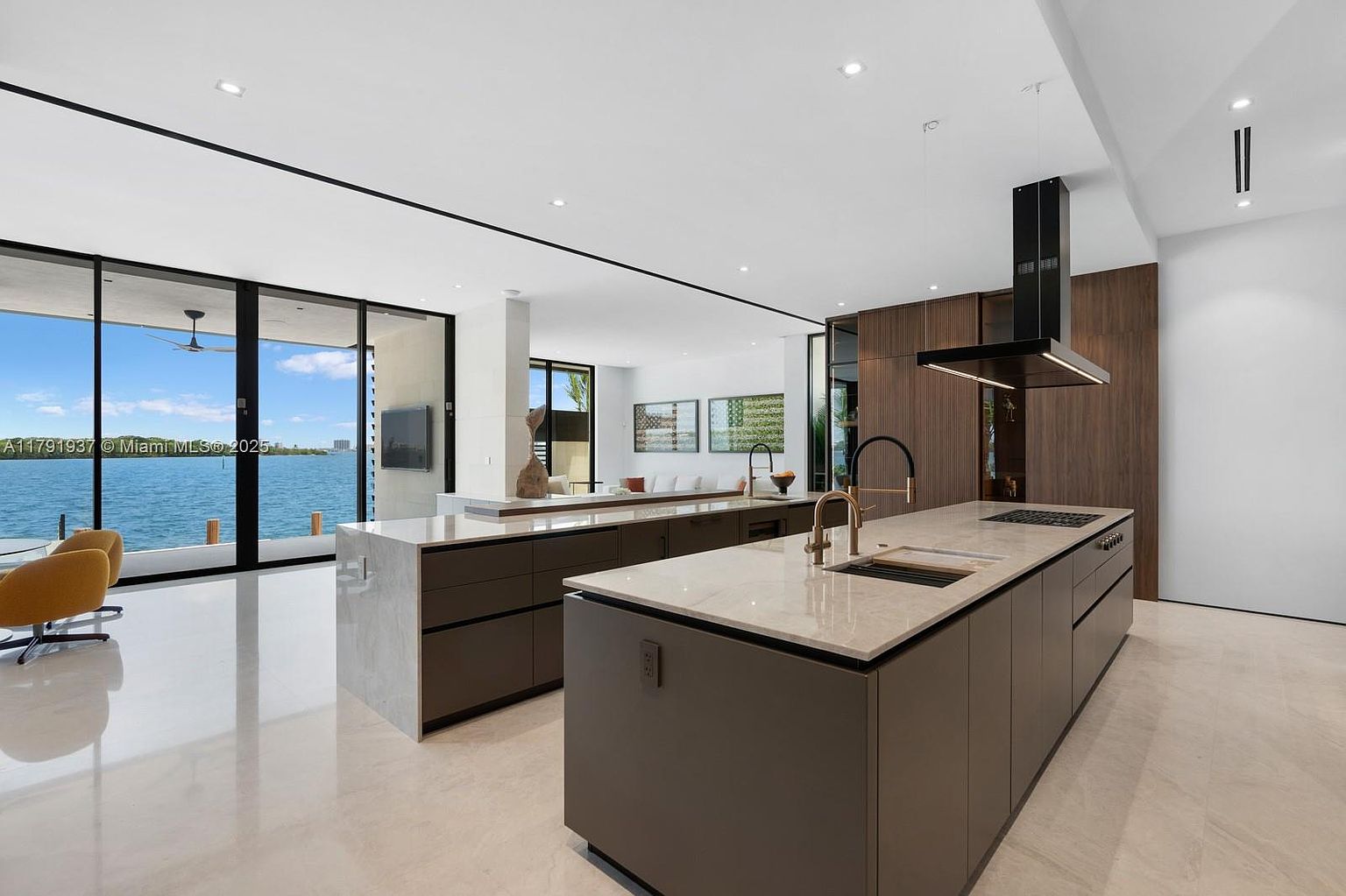 This is a modern, open-concept kitchen featuring two large islands with marble countertops and dark cabinetry. The kitchen is connected to a living area with floor-to-ceiling windows offering waterfront views. A sleek black range hood hangs above the cooktop, adding a touch of sophistication.