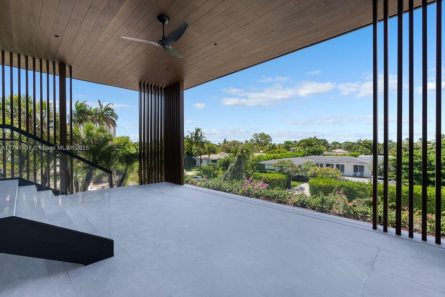 This image showcases a modern balcony or patio area with sleek, light-colored flooring and a contemporary design. The space features vertical wooden slats that provide both privacy and architectural interest, framing a view of lush greenery and neighboring houses. A ceiling fan adds to the comfort of the outdoor space, suggesting a relaxing and stylish area for enjoying the surrounding landscape.