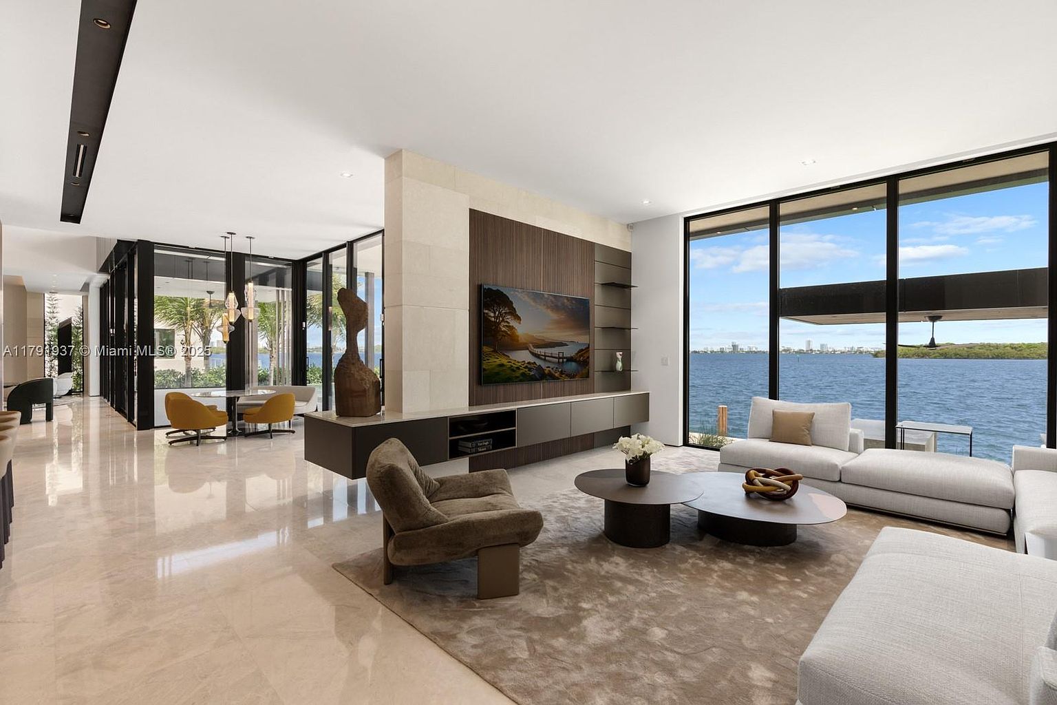 This is a spacious and modern living room with floor-to-ceiling windows offering a stunning water view. The room features a neutral color palette, contemporary furniture, and a large area rug. A sleek entertainment center with a mounted TV divides the living area from the dining space, creating an open and airy feel.