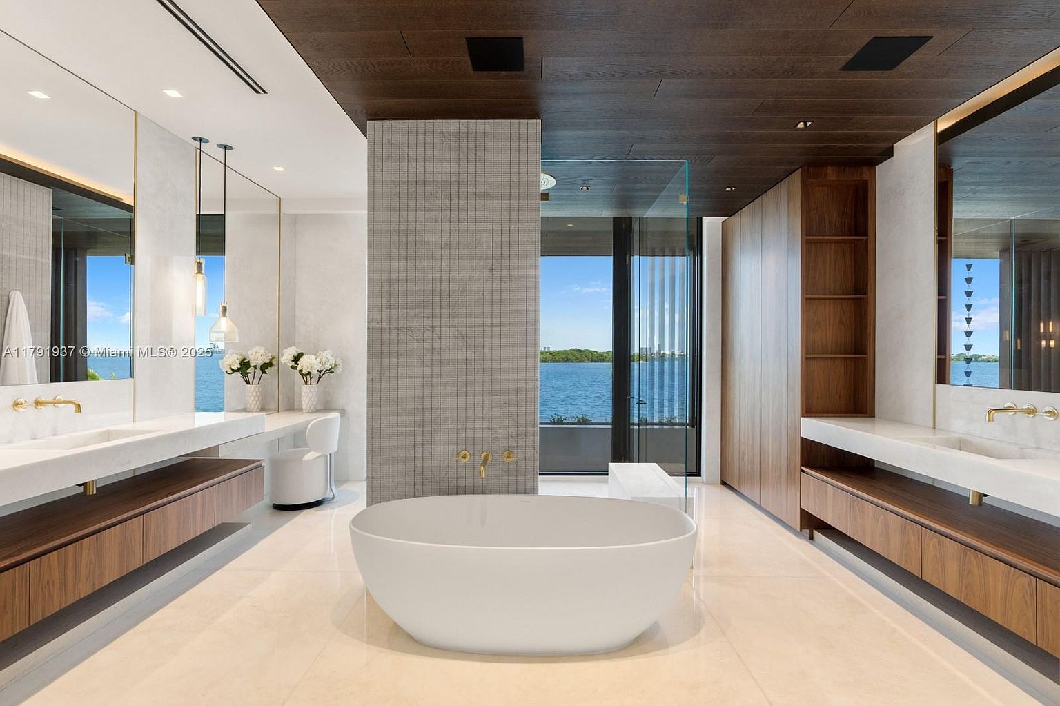 This is a luxurious primary bathroom featuring a freestanding white bathtub as a focal point. The room boasts dual vanities with modern fixtures, wood cabinetry, and a large window offering a scenic water view. The design incorporates a neutral color palette with wood accents, creating a serene and spa-like atmosphere.