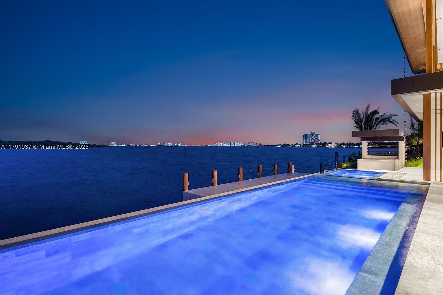 This image showcases a luxurious infinity pool overlooking a serene body of water, likely an ocean or bay, with a cityscape visible in the distance. The pool's blue lighting creates a tranquil ambiance, complemented by the modern architectural design of the adjacent building, which features clean lines and natural wood accents. The scene evokes a sense of high-end living and relaxation.