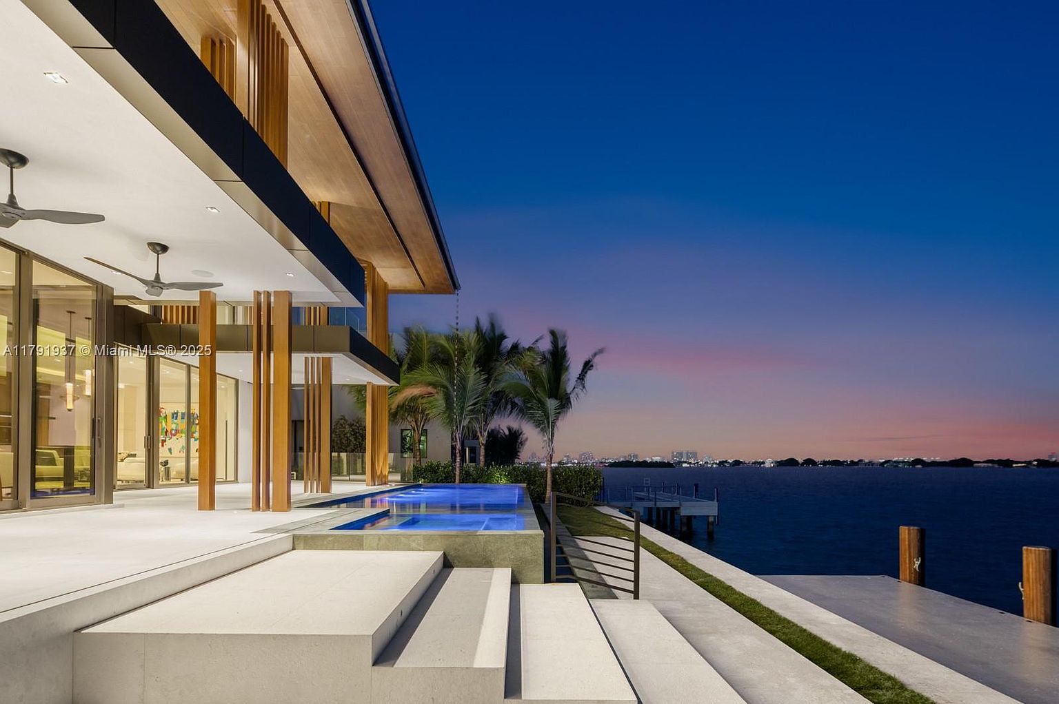 This image showcases the exterior rear view of a modern luxury home at dusk, featuring a sleek pool with illuminated water, a multi-level patio with steps leading down to the waterfront, and palm trees adding to the tropical ambiance. The architecture is contemporary with clean lines, and the overall impression is one of high-end waterfront living.