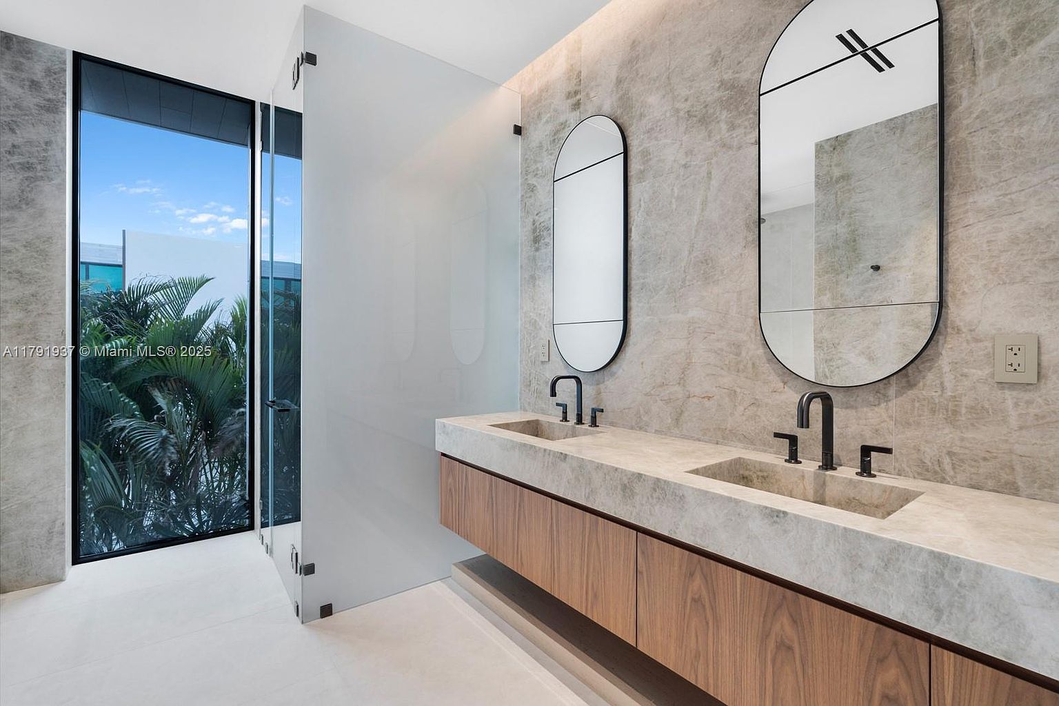 This is a modern primary bathroom featuring a double vanity with a light-colored stone countertop and wood cabinetry. Two oval mirrors with black frames hang above the sinks, complemented by sleek black faucets. A glass-enclosed shower area is visible to the left, offering a view of lush greenery outside the window, creating a spa-like atmosphere.
