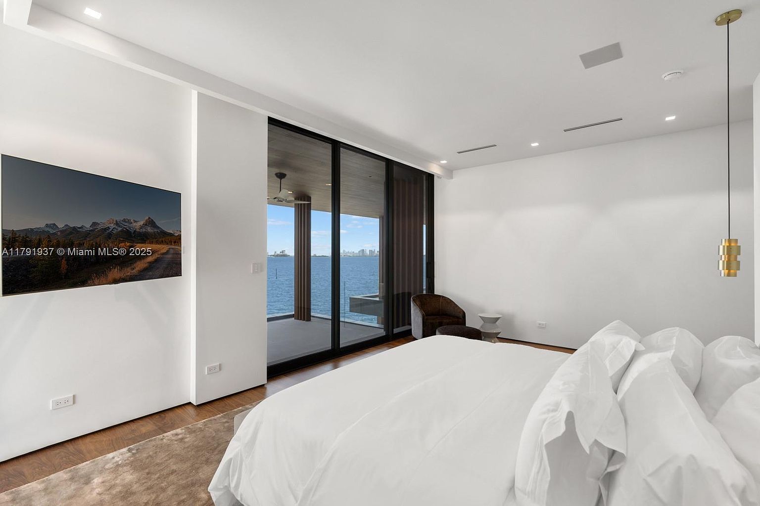 This is a bright and airy primary bedroom featuring a large bed with white linens, a modern pendant light, and a wall-mounted television. A sliding glass door leads to a balcony with ocean views, enhancing the room's luxurious feel. The room is minimalist in design with white walls and hardwood floors.