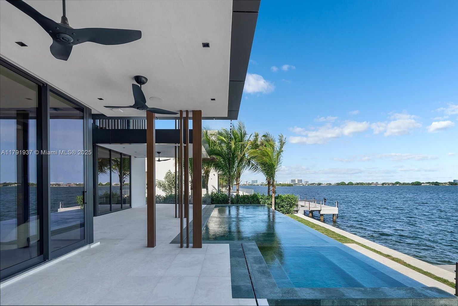 This image showcases a luxurious infinity pool extending towards a serene waterfront. The modern architectural design features clean lines, with dark-framed glass doors and wooden support beams adding a touch of elegance. The pool's reflective surface mirrors the sky, creating a seamless blend with the surrounding natural beauty.