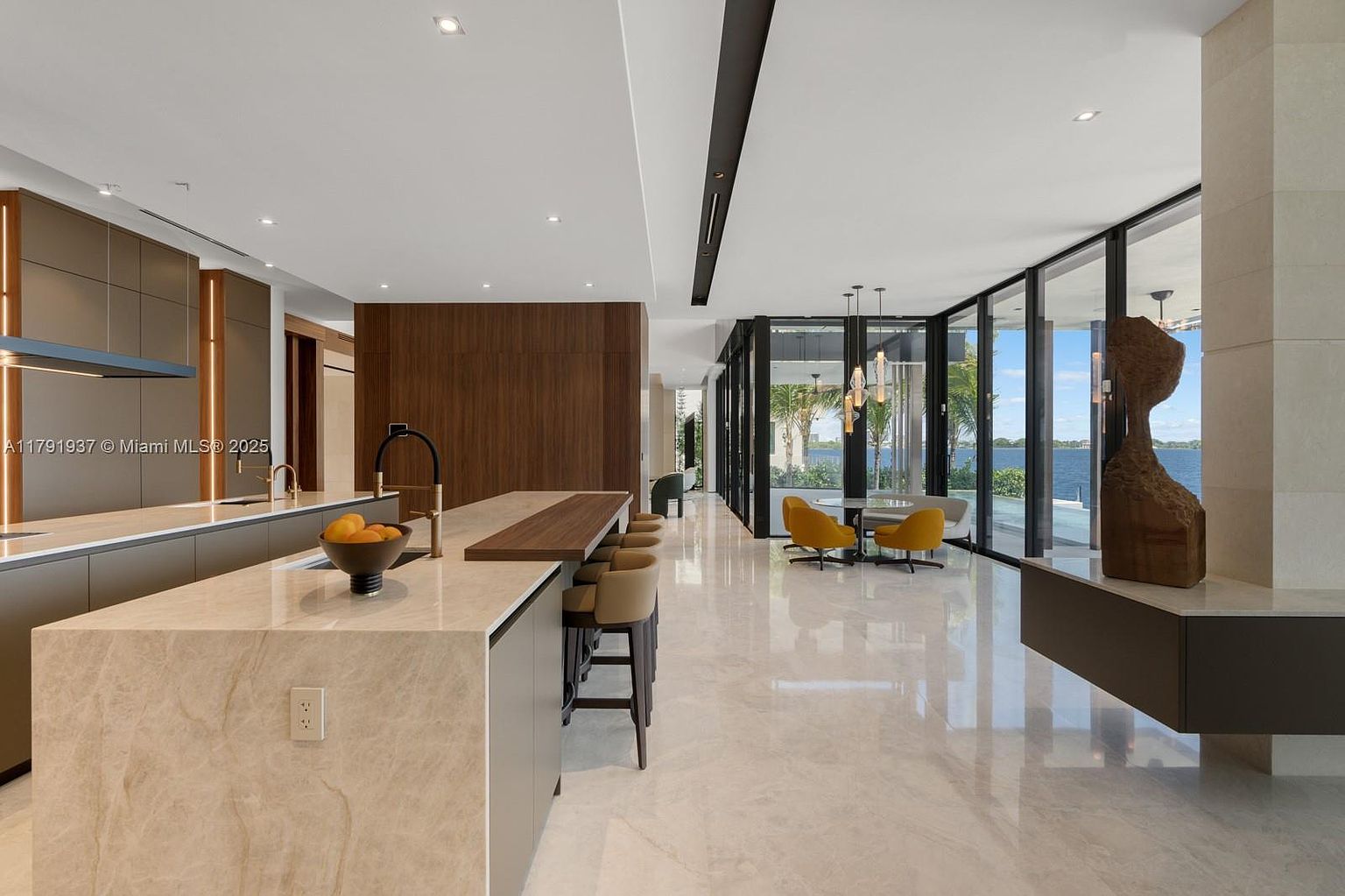 This interior shot showcases a modern, open-concept kitchen and dining area. The kitchen features a large island with a light-colored countertop and dark wood accents, complemented by sleek cabinetry and high-end appliances. The dining area, visible through expansive glass doors, offers a view of the water, creating a luxurious and inviting atmosphere.