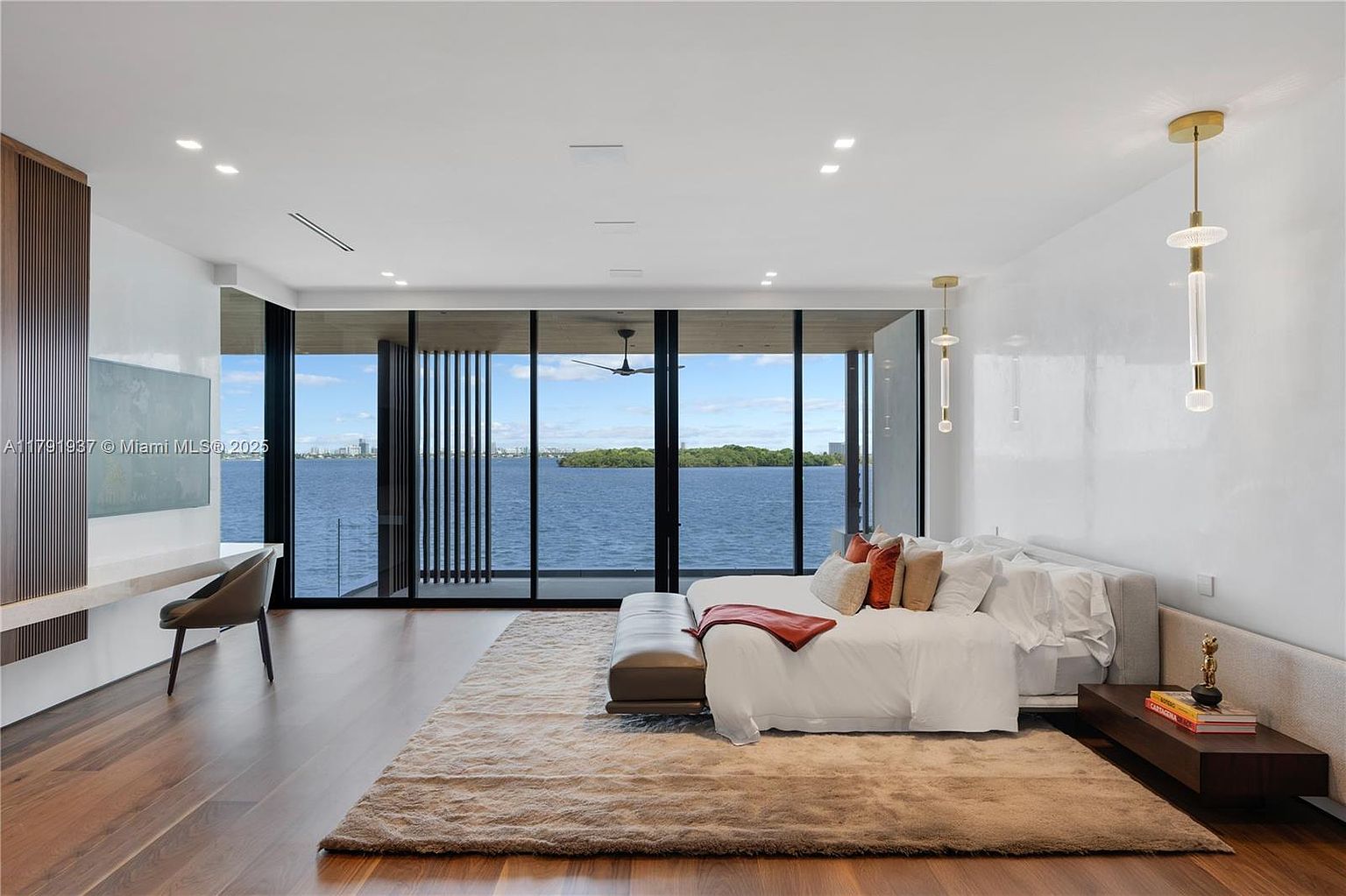 This is a luxurious primary bedroom featuring floor-to-ceiling windows with a stunning water view. The room is decorated in a modern style with a neutral color palette, hardwood floors, and a plush area rug. A sleek desk area and a comfortable bed with high-end linens add to the room's appeal, creating a serene and upscale atmosphere.