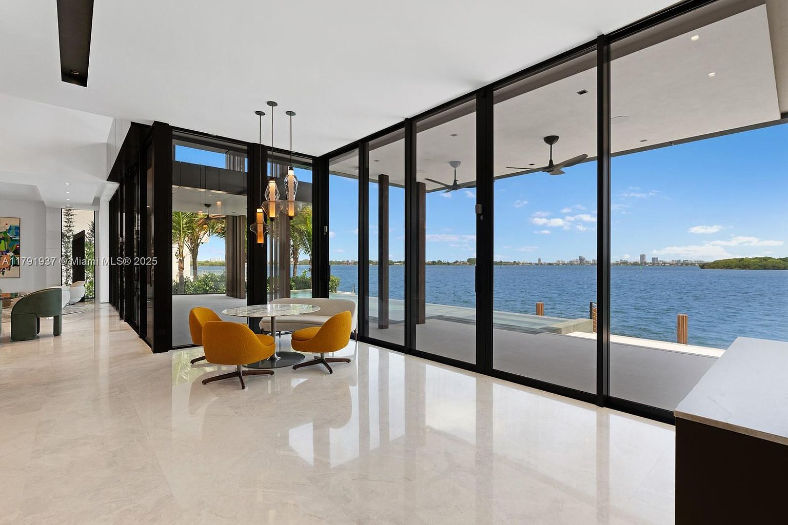 This is a bright and modern living room featuring floor-to-ceiling windows with a waterfront view. The room is furnished with a round table and yellow chairs, complemented by a sleek lighting fixture. The polished marble floors reflect the natural light, enhancing the spacious and luxurious feel of the room.