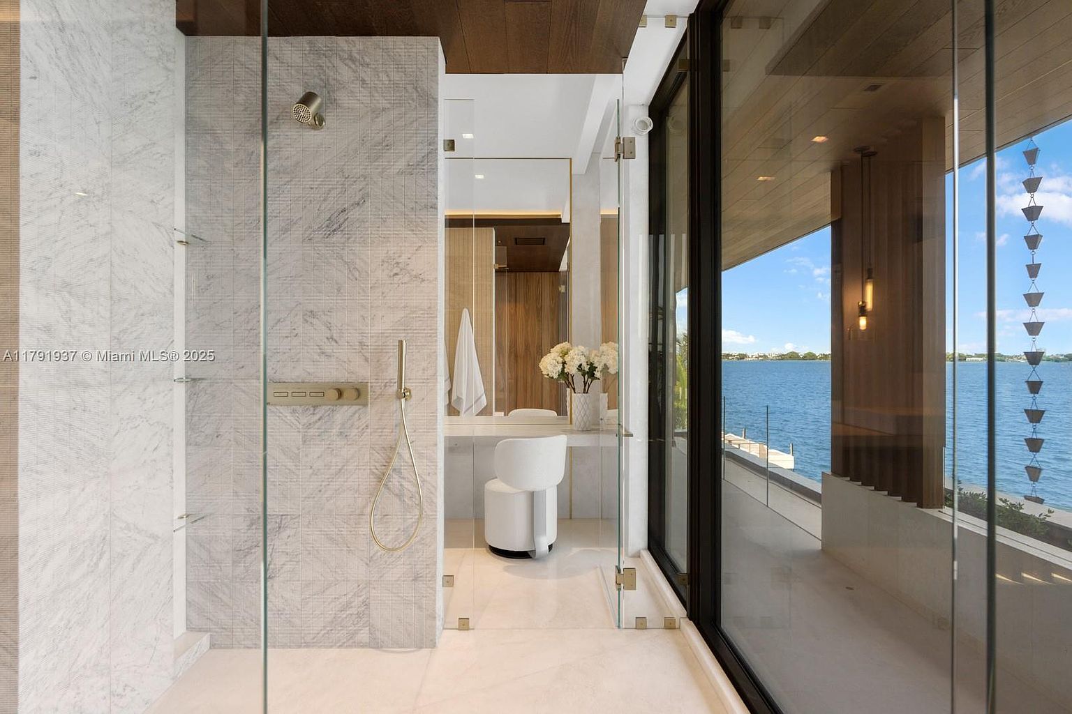 This is a luxurious primary bathroom featuring a glass-enclosed shower with marble tile and gold fixtures. The vanity area includes a modern chair, a large mirror, and fresh flowers. Large windows offer stunning water views, enhancing the spa-like atmosphere and highlighting the property's waterfront location.