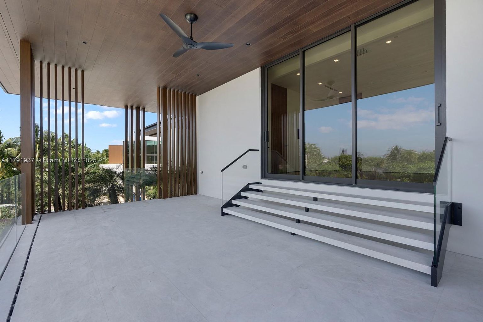 This image showcases a modern balcony or deck area with a sleek design. The space features a wooden ceiling with a ceiling fan, vertical wooden slats for privacy, and a set of stairs leading to a sliding glass door. The flooring is a light gray, and the overall aesthetic is clean and contemporary, offering a luxurious outdoor living space.