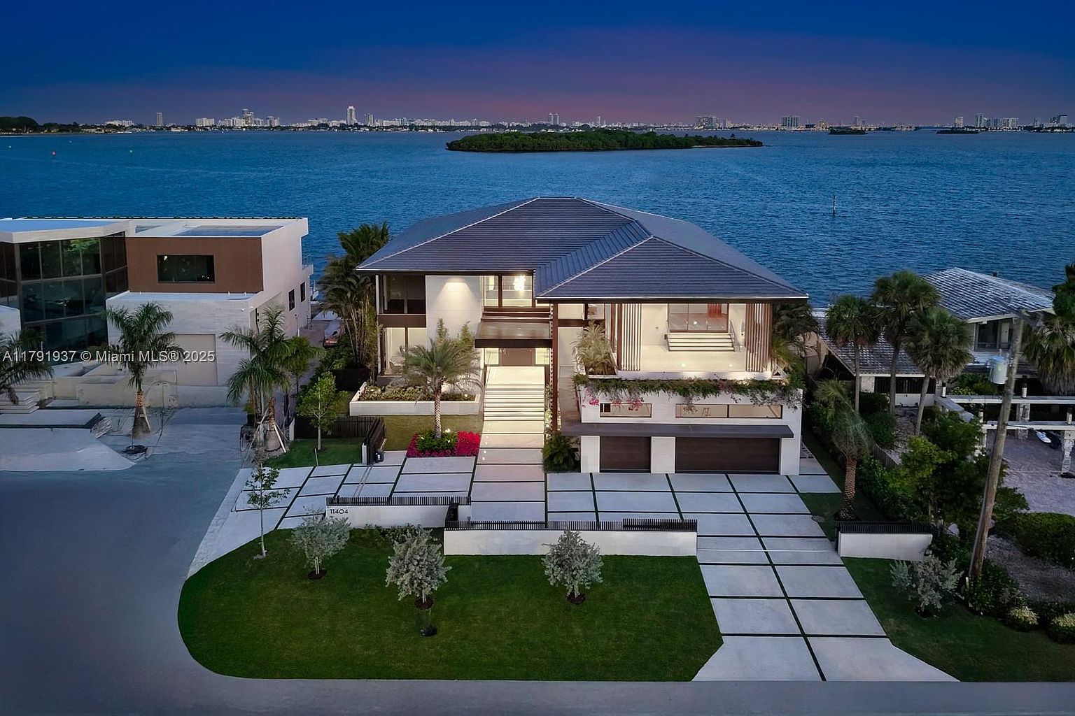 This is a stunning front view of a modern waterfront home. The property features a meticulously landscaped yard with geometric concrete pathways leading to the entrance. The house boasts a multi-level design with large windows, a gray roof, and a grand staircase leading to the main entrance, all set against a backdrop of a serene bay and distant city skyline.