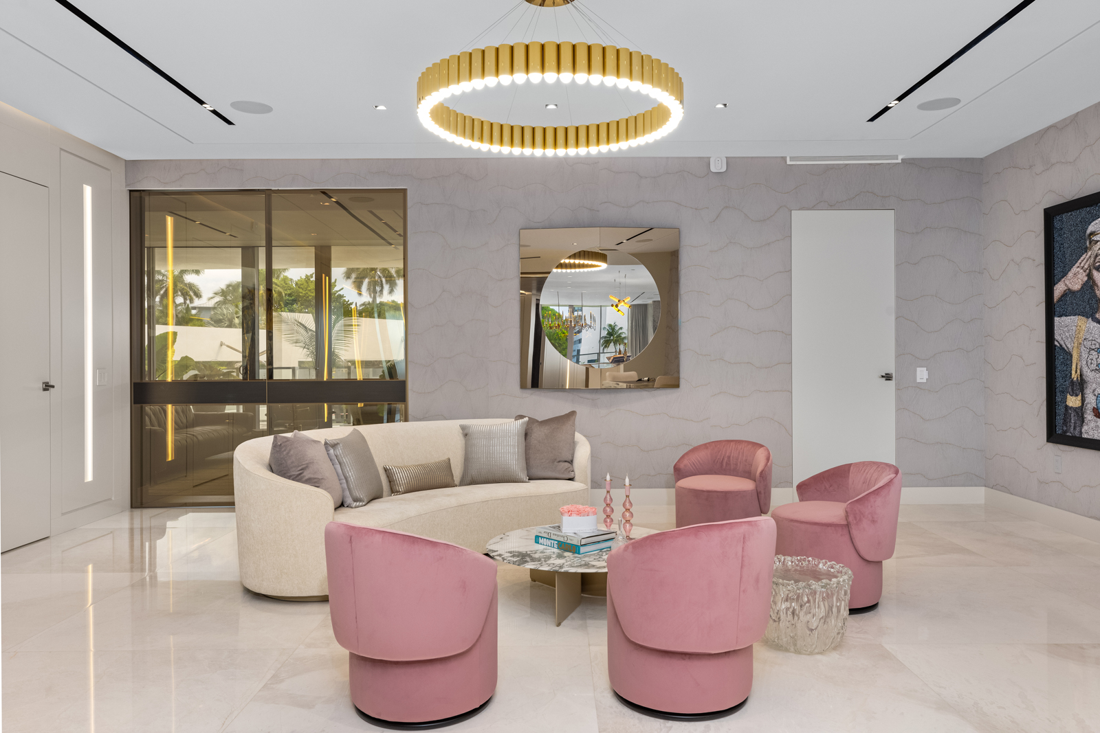 This interior shot showcases a stylish living room with a curved sofa, pink armchairs, and a marble-topped coffee table. A modern, circular chandelier hangs above, and a large mirror reflects the room's design. The space is bright and airy, with a sophisticated and inviting atmosphere.