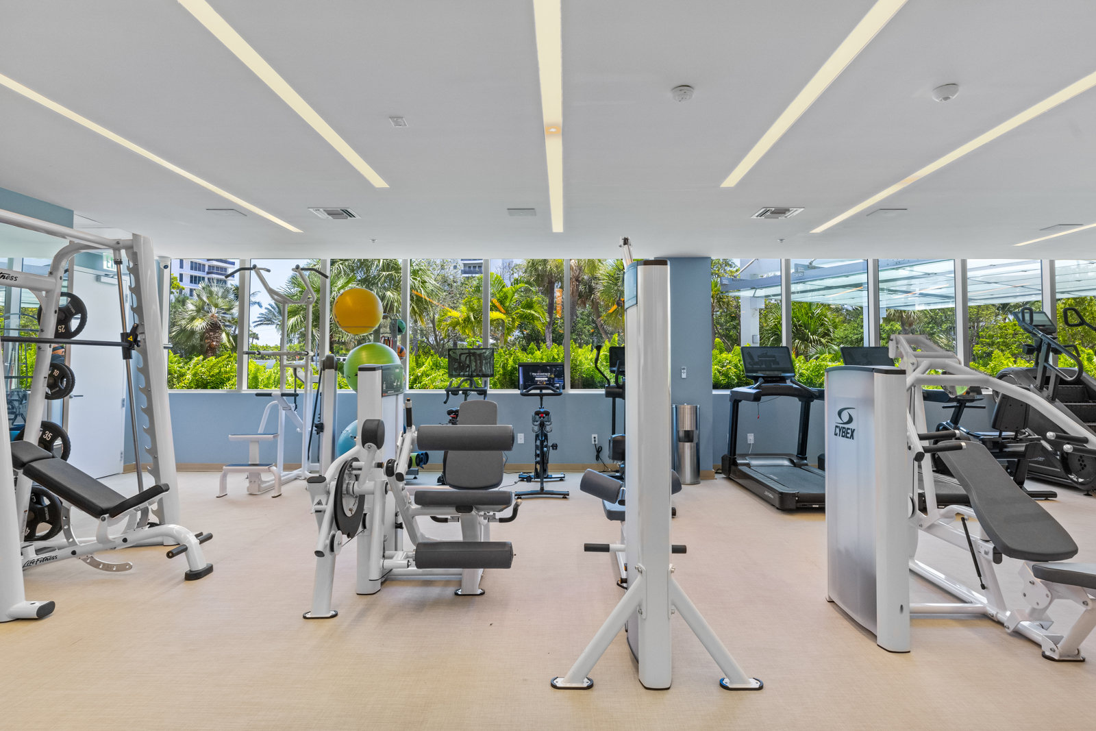 This is a well-equipped home gym featuring various exercise machines, including weightlifting stations, treadmills, and stationary bikes. Large windows provide ample natural light and views of lush greenery outside. The space is clean, modern, and designed for a comprehensive workout experience.