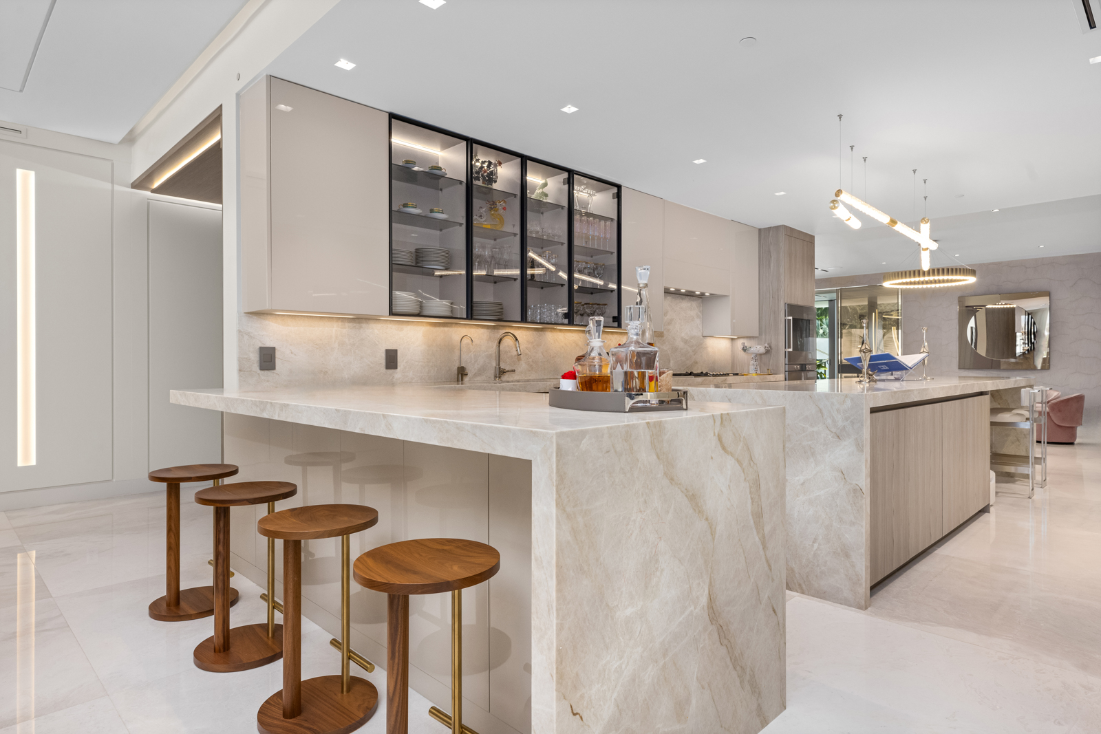 This is a bright and modern kitchen featuring a large island with a marble countertop and waterfall edges, complemented by wooden bar stools with gold accents. The kitchen includes sleek, light-colored cabinetry, glass-fronted display cabinets, and stainless steel appliances. The open layout and elegant lighting fixtures create a luxurious and inviting space.
