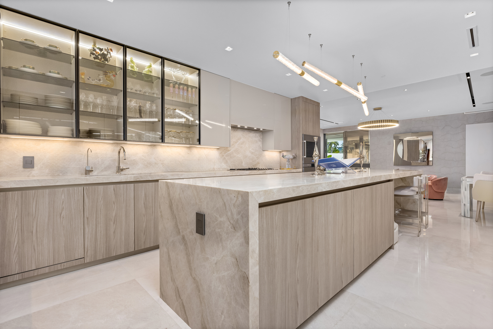 This is a bright and modern kitchen featuring light wood-grain cabinetry, marble countertops, and a large island. Glass-front cabinets display dishware and glassware, while contemporary pendant lighting illuminates the space. The kitchen seamlessly transitions into a dining area, creating an open and inviting atmosphere.