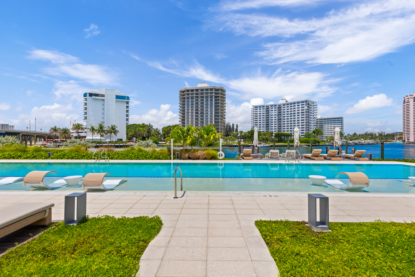 This image showcases a luxurious outdoor pool area with modern lounge chairs partially submerged in the water, offering a unique relaxation experience. The pool is surrounded by well-maintained landscaping and a tiled patio, with high-rise buildings visible in the background, suggesting a prime waterfront location. The scene evokes a sense of upscale resort living and relaxation.
