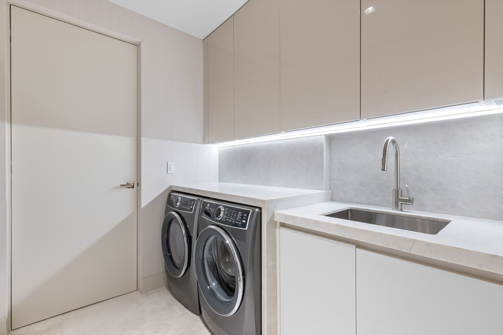 This is a well-lit, modern laundry room featuring a front-loading washer and dryer set in a dark gray finish. The room includes light-colored cabinetry above and below a countertop, a stainless steel sink with a modern faucet, and a door on the left. The overall impression is clean, organized, and functional, perfect for a modern home.