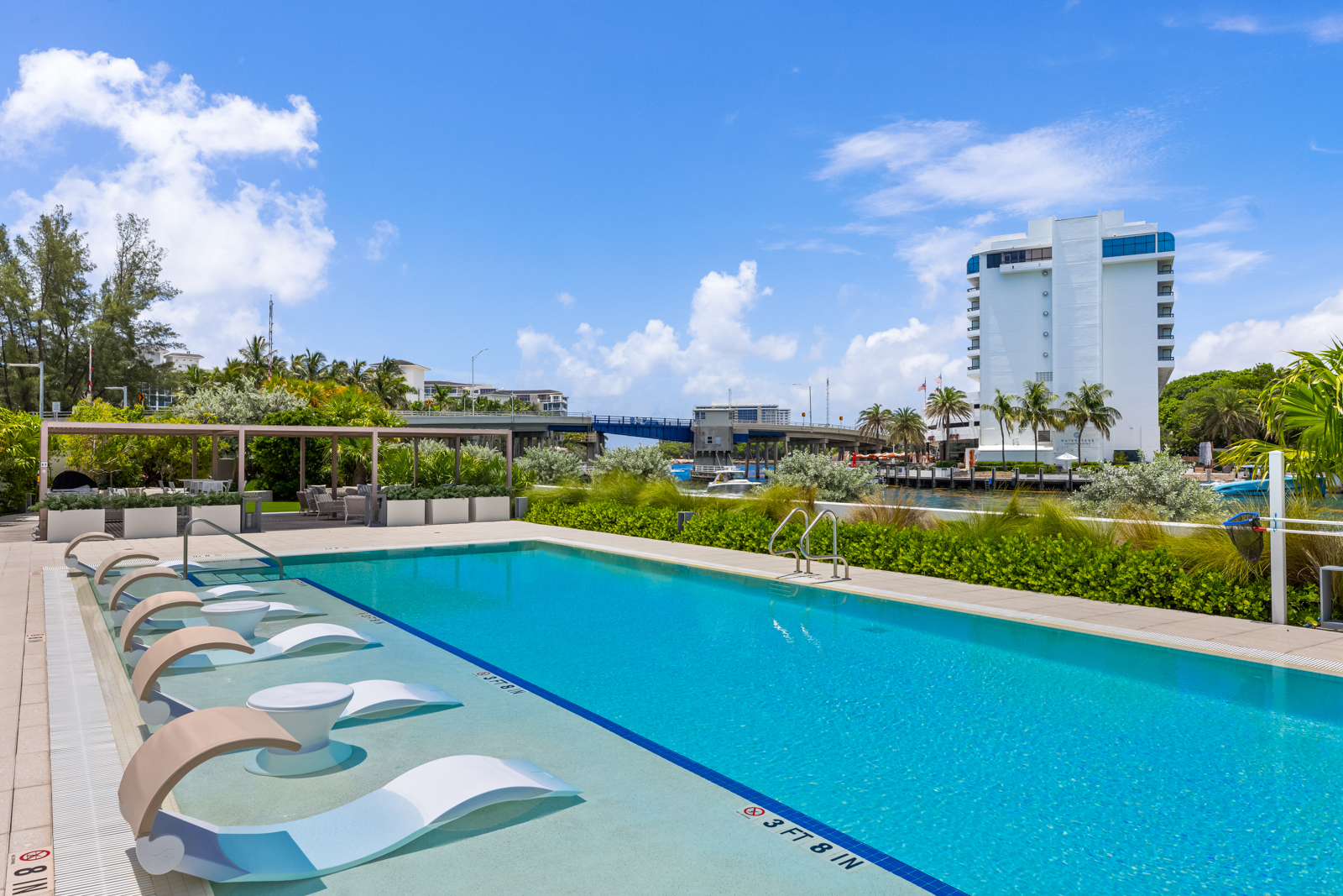 This image showcases a luxurious outdoor pool area, featuring in-water lounge chairs and a clear blue pool. Lush greenery surrounds the pool, providing privacy and a tropical feel, while a modern building and a bridge are visible in the background. The scene evokes a sense of relaxation and upscale living.