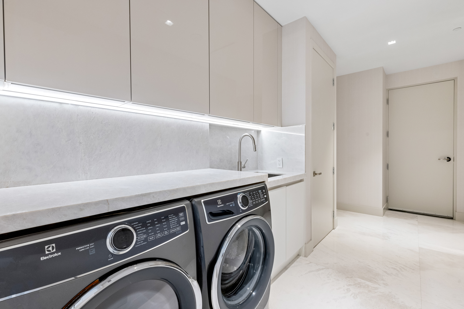 This is a modern laundry room featuring sleek, light-colored cabinetry and countertops. A front-loading washer and dryer set is prominently displayed, suggesting convenience and efficiency. The room is well-lit and appears clean, creating a functional and aesthetically pleasing space.