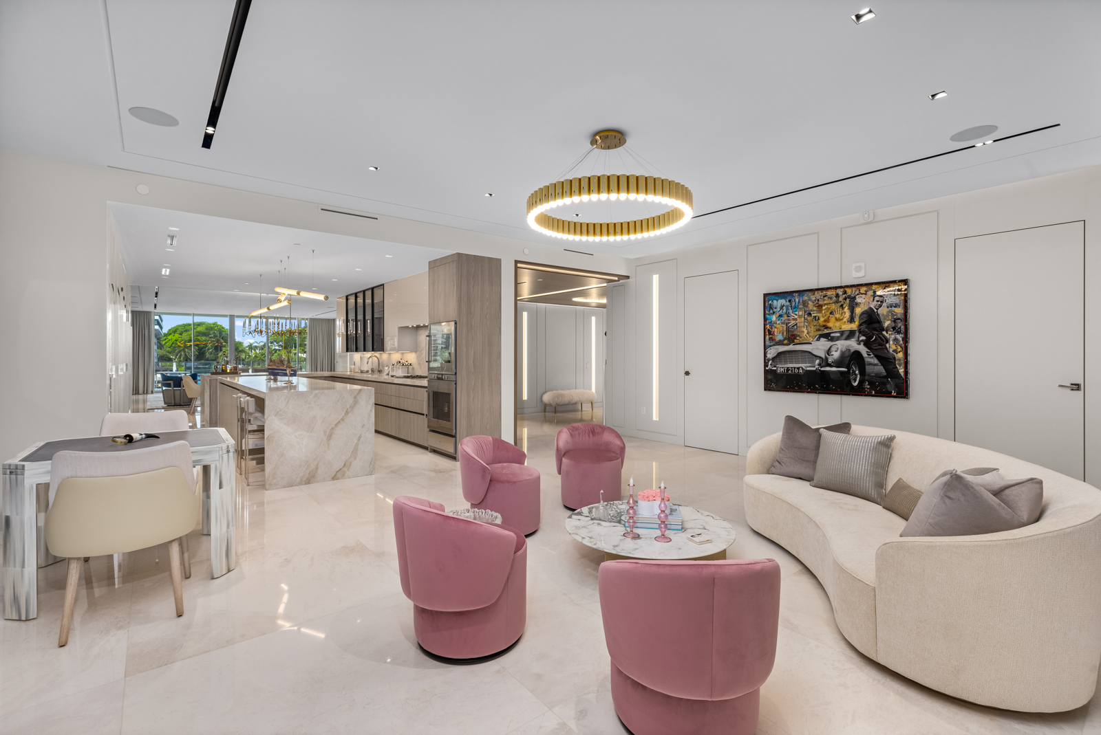 This interior shot showcases a luxurious living room with a modern aesthetic. The room features a curved sofa, four pink velvet chairs arranged around a marble coffee table, and a striking circular chandelier. The open floor plan seamlessly connects to the kitchen and dining area, enhancing the sense of spaciousness and flow.