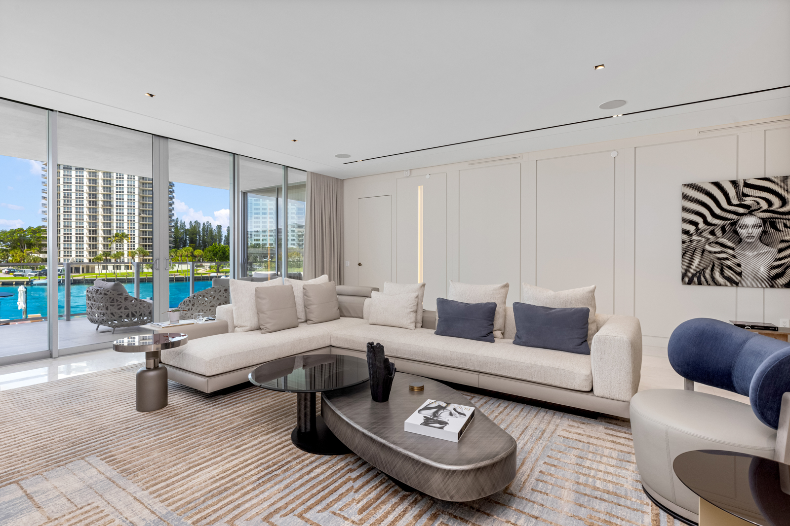 This is a bright and modern living room featuring a large sectional sofa with neutral-toned cushions and blue accent pillows. A unique coffee table sits atop a patterned rug, and floor-to-ceiling windows offer a view of the waterfront. The room is decorated with contemporary art and stylish furniture, creating a luxurious and inviting atmosphere.