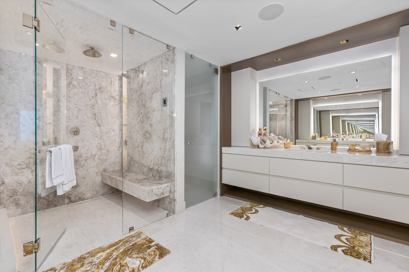 This is a luxurious primary bathroom featuring a spacious walk-in shower with marble walls and a glass enclosure. The vanity area includes a large mirror with integrated lighting, white cabinetry, and elegant countertops. The flooring is light-colored tile, and decorative rugs add a touch of warmth to the space.