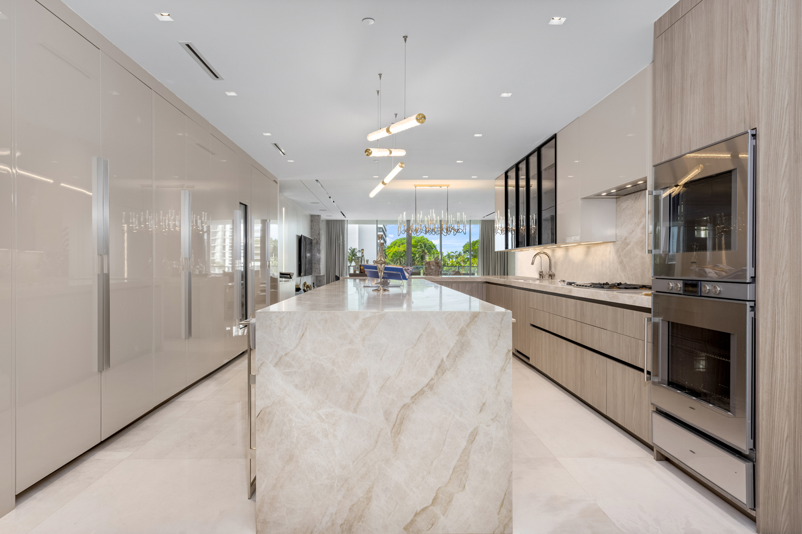 This is a bright, modern kitchen featuring sleek, glossy cabinetry on one side and wood-paneled cabinets on the other. A large marble island takes center stage, illuminated by contemporary pendant lighting. Stainless steel appliances and a view through a large window enhance the luxurious feel of the space.