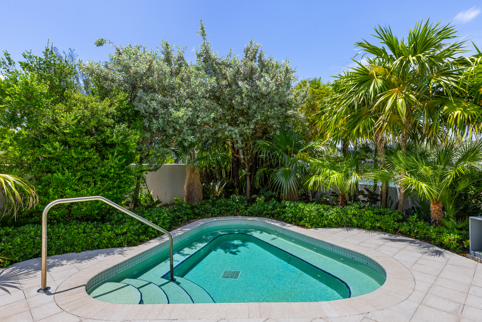 This image showcases a luxurious spa surrounded by lush greenery, including palm trees and various shrubs, creating a private and serene oasis. The spa features a unique triangular shape with tiled steps and a stainless steel handrail for easy access. The surrounding patio is paved with light gray stones, enhancing the upscale and tranquil ambiance.