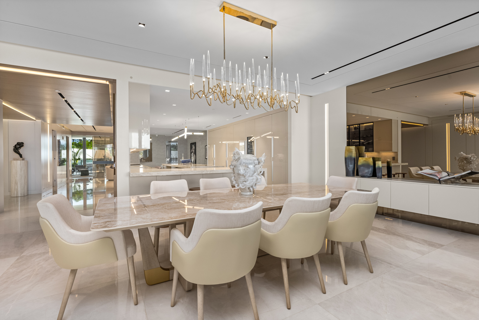 This interior shot showcases a luxurious dining room featuring a marble-topped dining table surrounded by elegant, upholstered chairs. A modern chandelier hangs above the table, providing ample lighting. The room's design is sophisticated and spacious, with a seamless transition to the adjacent kitchen area, creating an open and inviting atmosphere.