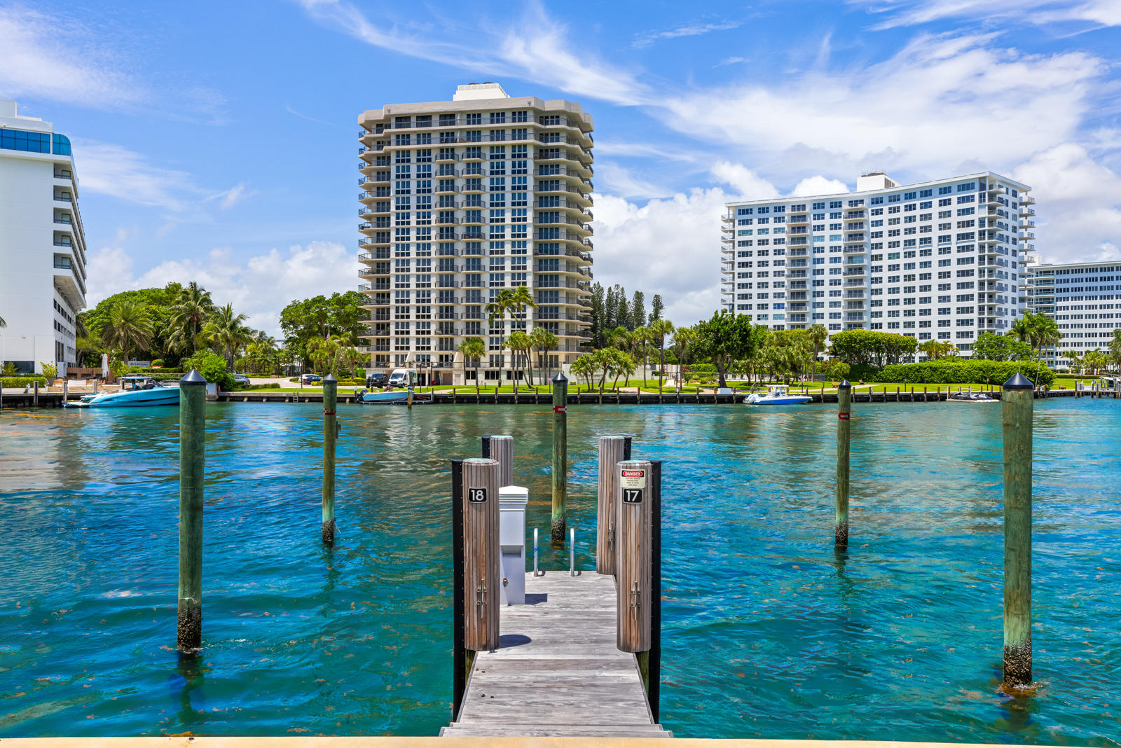 This image showcases a waterfront view with a wooden dock in the foreground, leading out into turquoise water. In the background, there are several high-rise residential buildings with white facades and numerous windows, suggesting luxury apartments or condominiums. Lush green trees and landscaping line the waterfront, adding to the serene and upscale atmosphere.