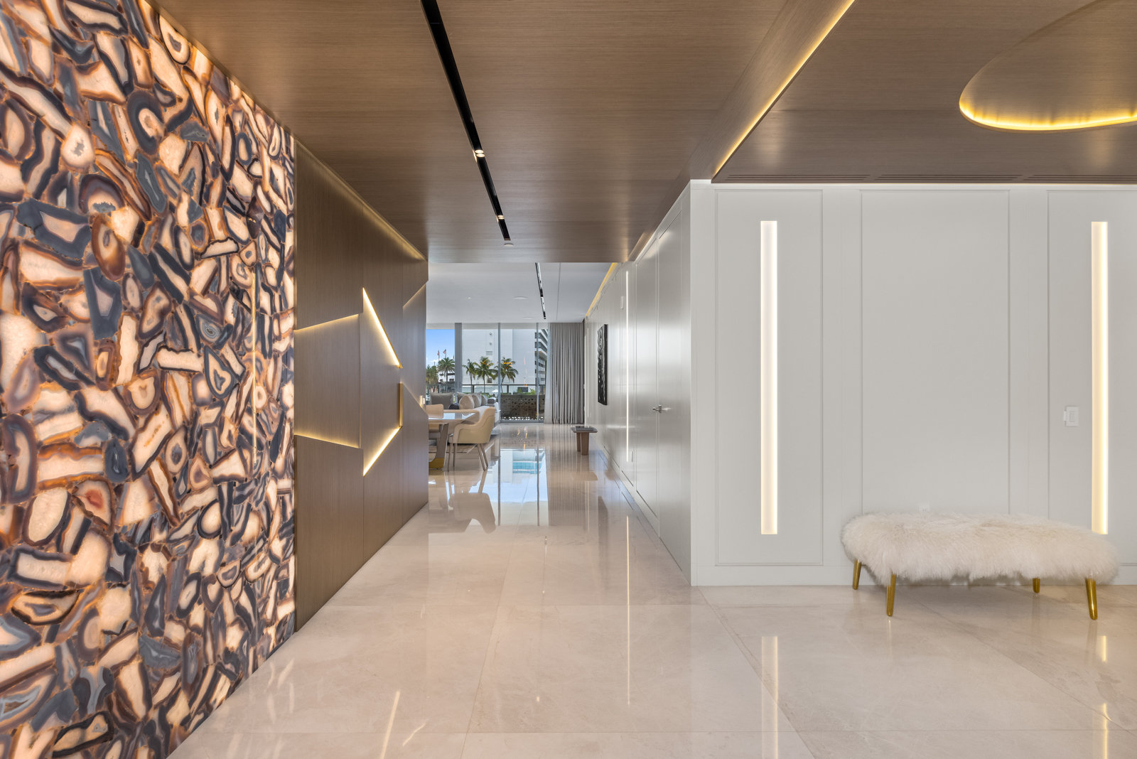 This interior shot showcases a luxurious hallway featuring a striking agate stone wall and sleek, modern design elements. The hallway is illuminated by recessed lighting and linear light fixtures, highlighting the polished marble flooring and creating a bright, open space. A plush bench with gold legs adds a touch of elegance and comfort.