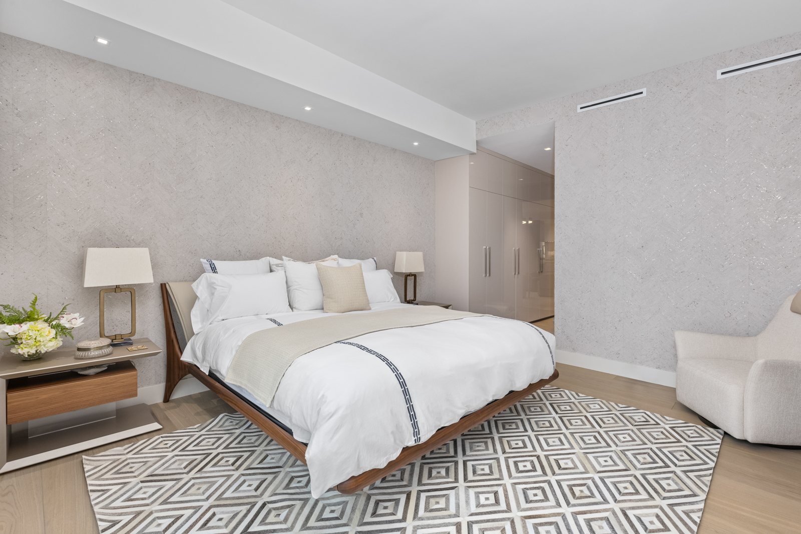 This is a well-lit primary bedroom featuring a modern platform bed with white linens and a decorative throw. The room is decorated with a geometric patterned rug, a side table with a lamp and floral arrangement, and a comfortable armchair. The walls are textured, and there's a built-in wardrobe in the background, creating a serene and stylish atmosphere.