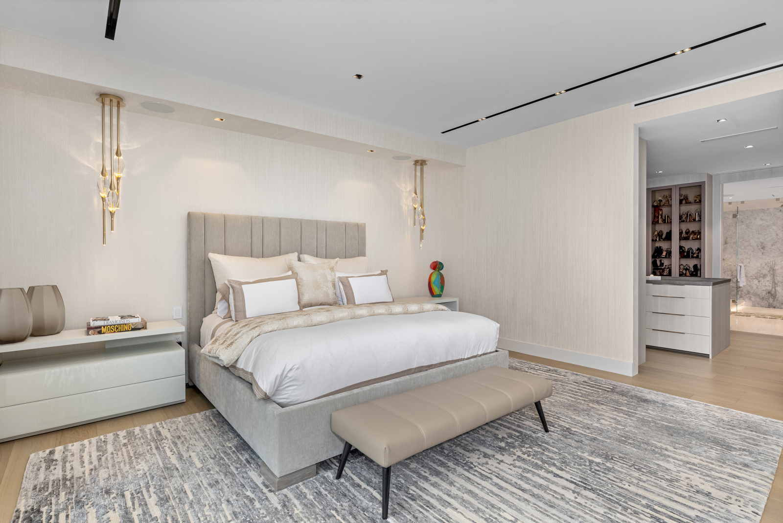 This is a well-lit primary bedroom featuring a large bed with a padded headboard, crisp white linens, and decorative pillows. The room is decorated in neutral tones with a textured wall covering, modern light fixtures, and a stylish area rug. A walk-in closet is visible in the background, adding to the room's appeal.