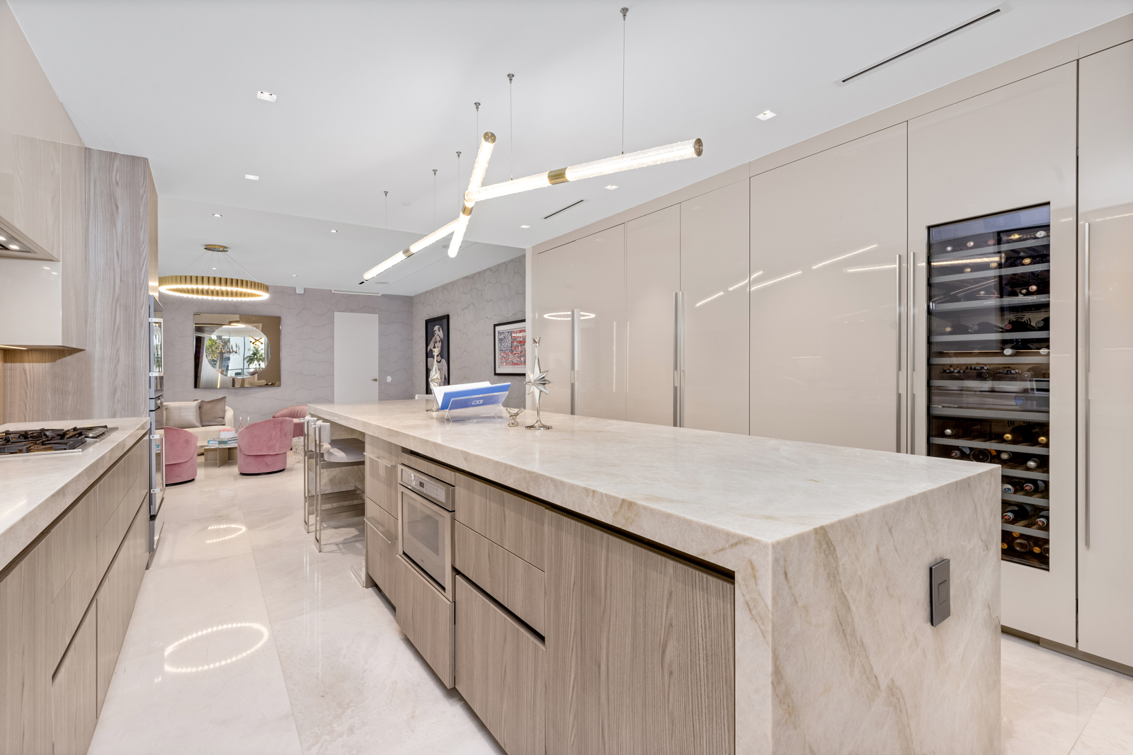 This is a modern, high-end kitchen featuring a large island with a marble countertop and wood-grain cabinetry. The kitchen includes built-in appliances, a wine refrigerator, and sleek, glossy cabinets. The open floor plan leads into a living area, creating a seamless and luxurious living space.