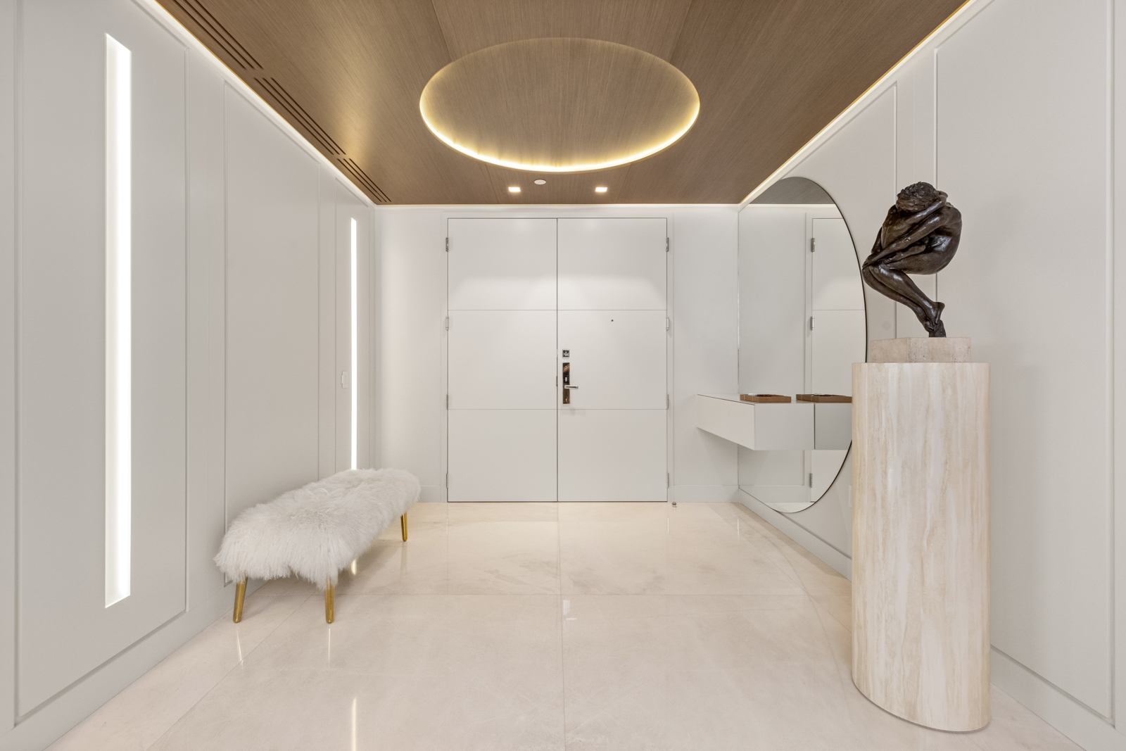 This is a well-lit, modern hallway featuring a clean, minimalist design. The walls are white with vertical recessed lighting, complemented by a light wood ceiling with a circular illuminated feature. A white faux fur bench sits to the left, and a sculpture on a travertine pedestal with a round mirror behind it is on the right, adding a touch of elegance.