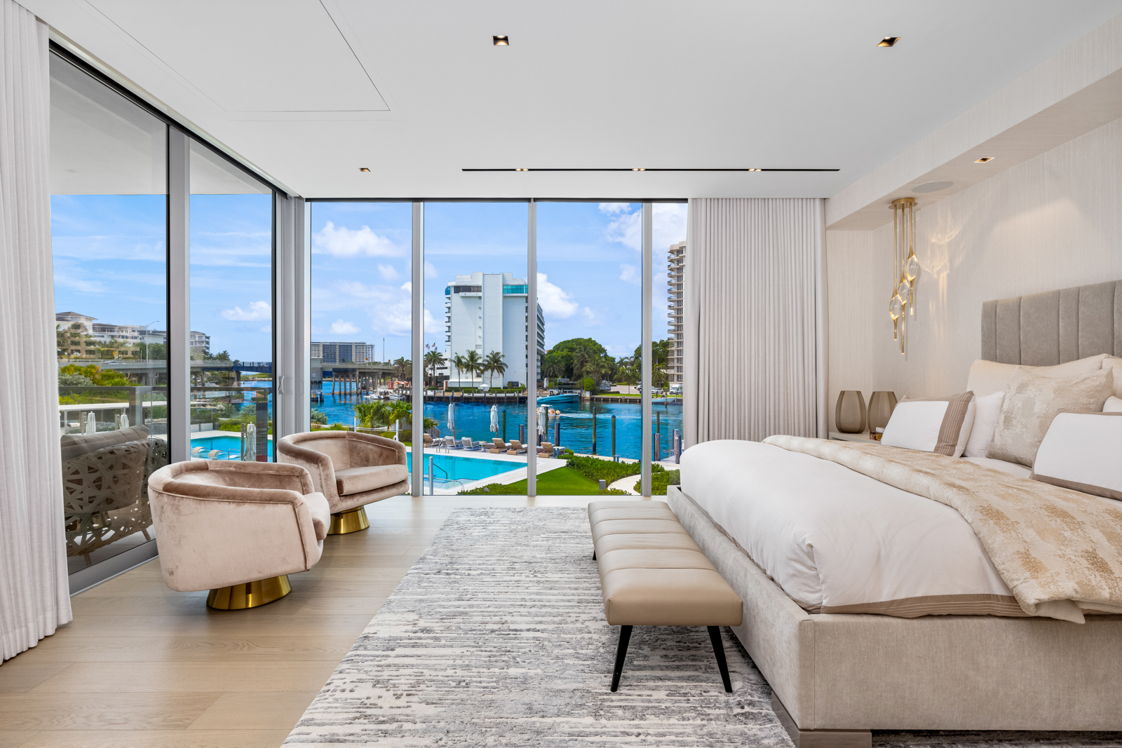 This is a luxurious primary bedroom featuring floor-to-ceiling windows with a waterfront view. The room is furnished with a king-size bed, a bench, and two swivel chairs. The color palette is neutral, creating a serene and elegant atmosphere, perfect for relaxation.