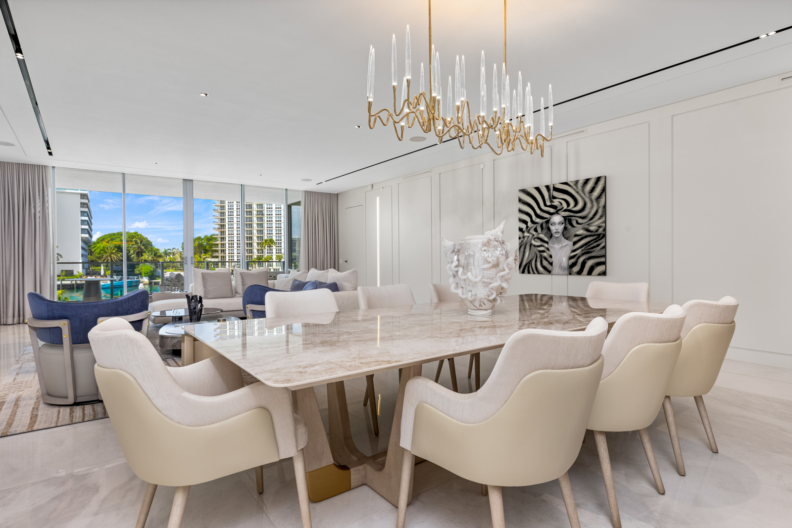 This interior shot showcases a luxurious dining room with a large marble-topped table surrounded by elegant beige chairs. A modern chandelier hangs above the table, and an abstract artwork adorns the wall. The room opens to a living area with views of a waterfront cityscape, creating a sophisticated and inviting atmosphere.
