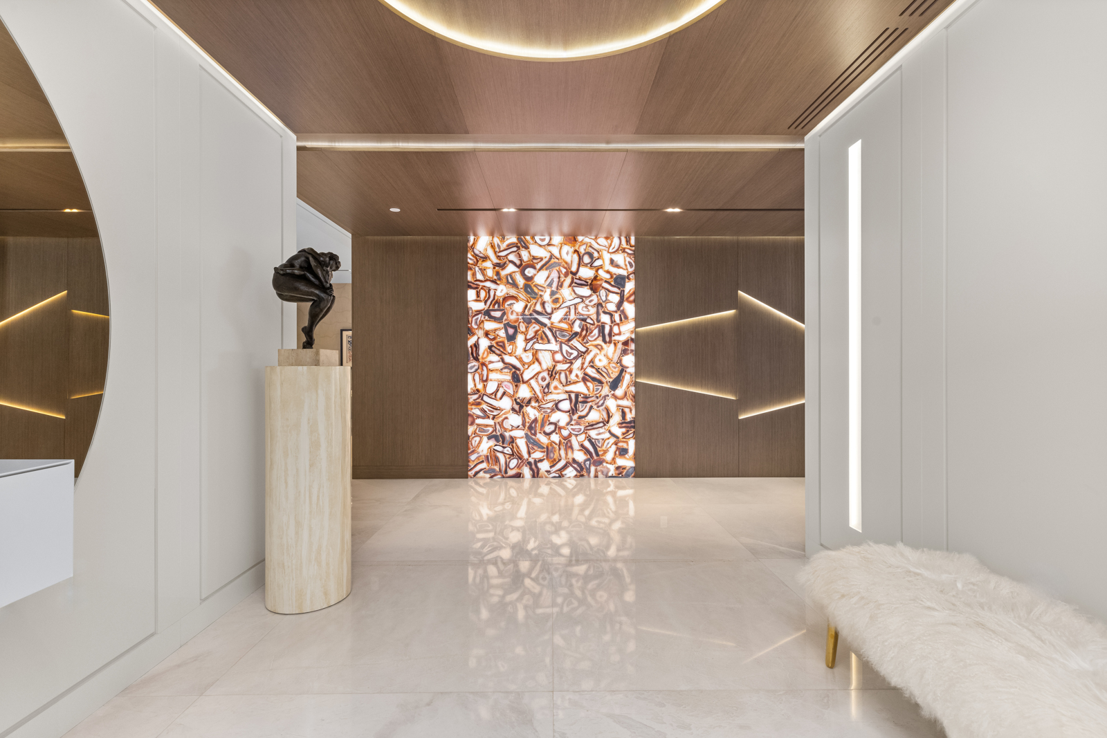 This interior shot showcases a luxurious hallway with a modern aesthetic. The space features a polished marble floor, wood-paneled walls with integrated lighting, and a striking backlit stone panel. A sculpture on a pedestal and a plush bench add to the sophisticated ambiance, creating an inviting and elegant entryway.