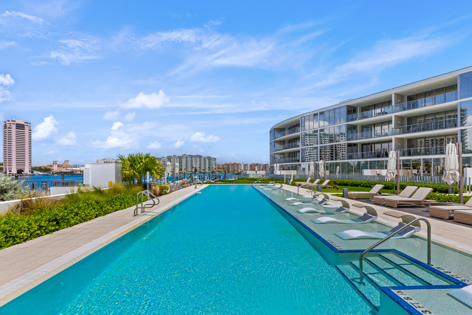 This image showcases a luxurious outdoor pool area with a stunning view of the city skyline and waterfront. The pool features a shallow lounging area with in-water chaise lounges, complemented by traditional lounge chairs and umbrellas on the deck. The modern architecture of the adjacent building adds to the upscale ambiance, making it an ideal amenity for a high-end property.
