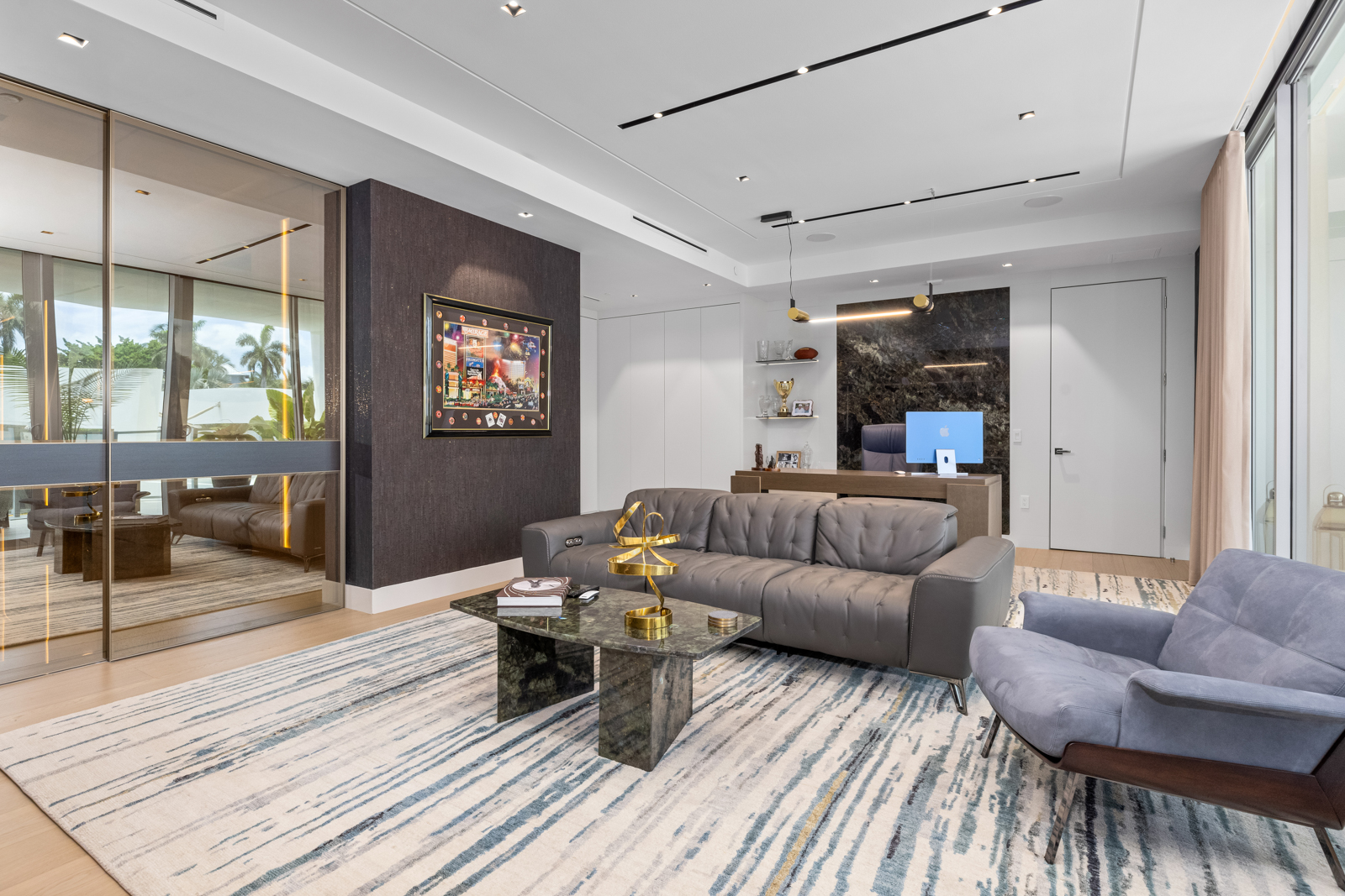 This is a modern office space featuring a gray leather sofa, a blue upholstered armchair, and a marble-topped coffee table on a patterned rug. The room includes a desk with a computer, built-in shelving, and a dark accent wall with artwork, creating a sophisticated and functional workspace. Large windows provide natural light, enhancing the contemporary design.