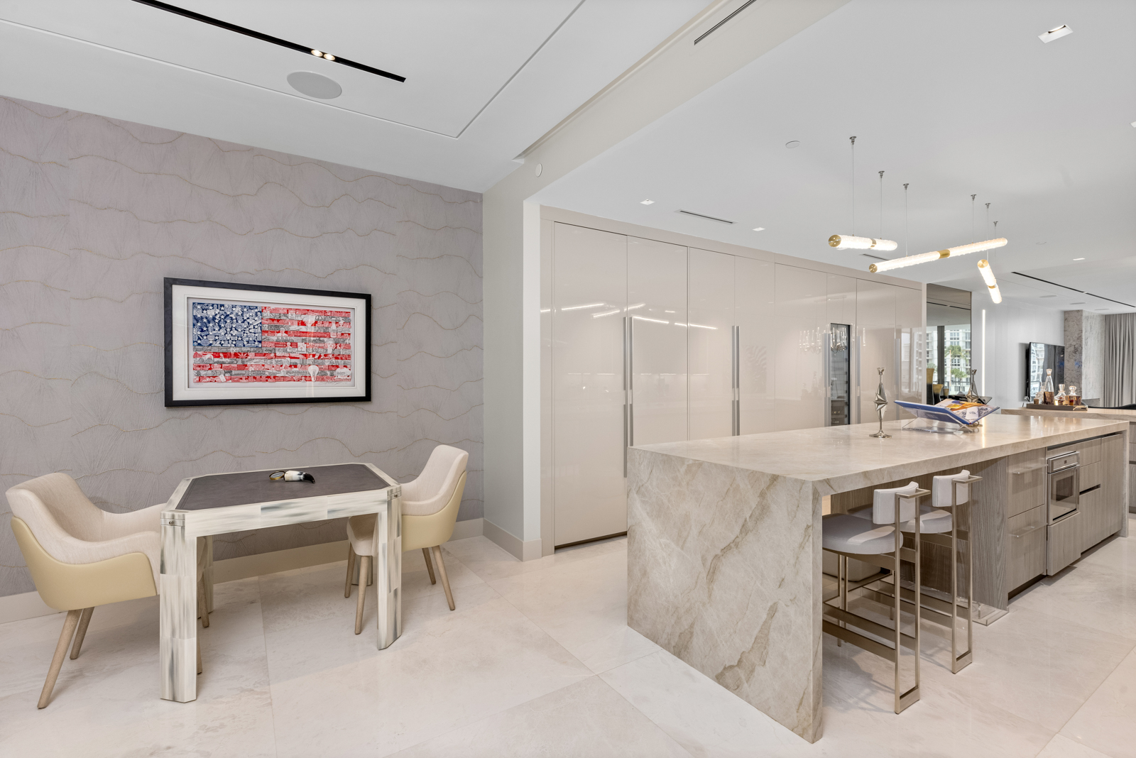 This interior shot showcases a modern kitchen and dining area. The kitchen features a large island with a marble countertop and bar seating, complemented by sleek cabinetry and modern lighting fixtures. Adjacent to the kitchen is a dining area with a small table and chairs, adorned with an American flag artwork, creating a cohesive and stylish living space.