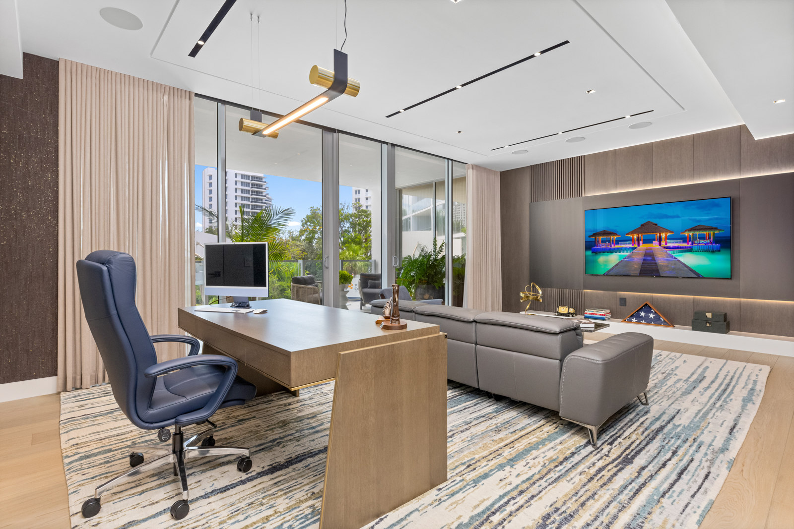 This is a modern and luxurious home office featuring a large wooden desk with a computer, a comfortable blue leather office chair, and a gray leather sofa. The room has large windows offering natural light and views of the outdoors, complemented by a stylish light fixture above the desk. A large television is mounted on the wall, and the room is decorated with tasteful accessories, creating a sophisticated and productive workspace.
