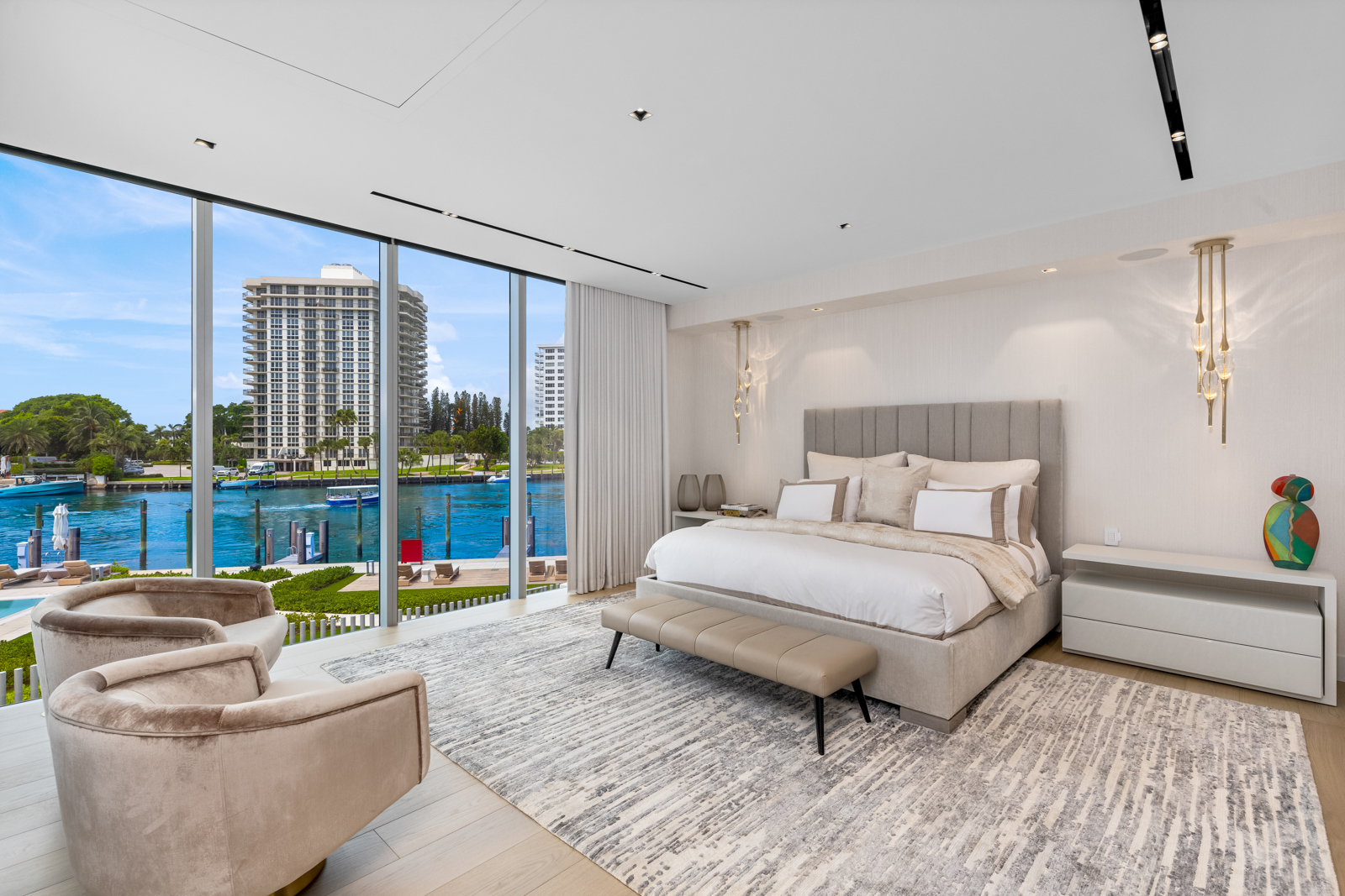This is a luxurious primary bedroom with a stunning waterfront view through floor-to-ceiling windows. The room features a neutral color palette, a large upholstered bed with multiple pillows, a bench at the foot of the bed, and a stylish area rug. Two modern armchairs are positioned to take advantage of the view, creating a serene and upscale atmosphere.