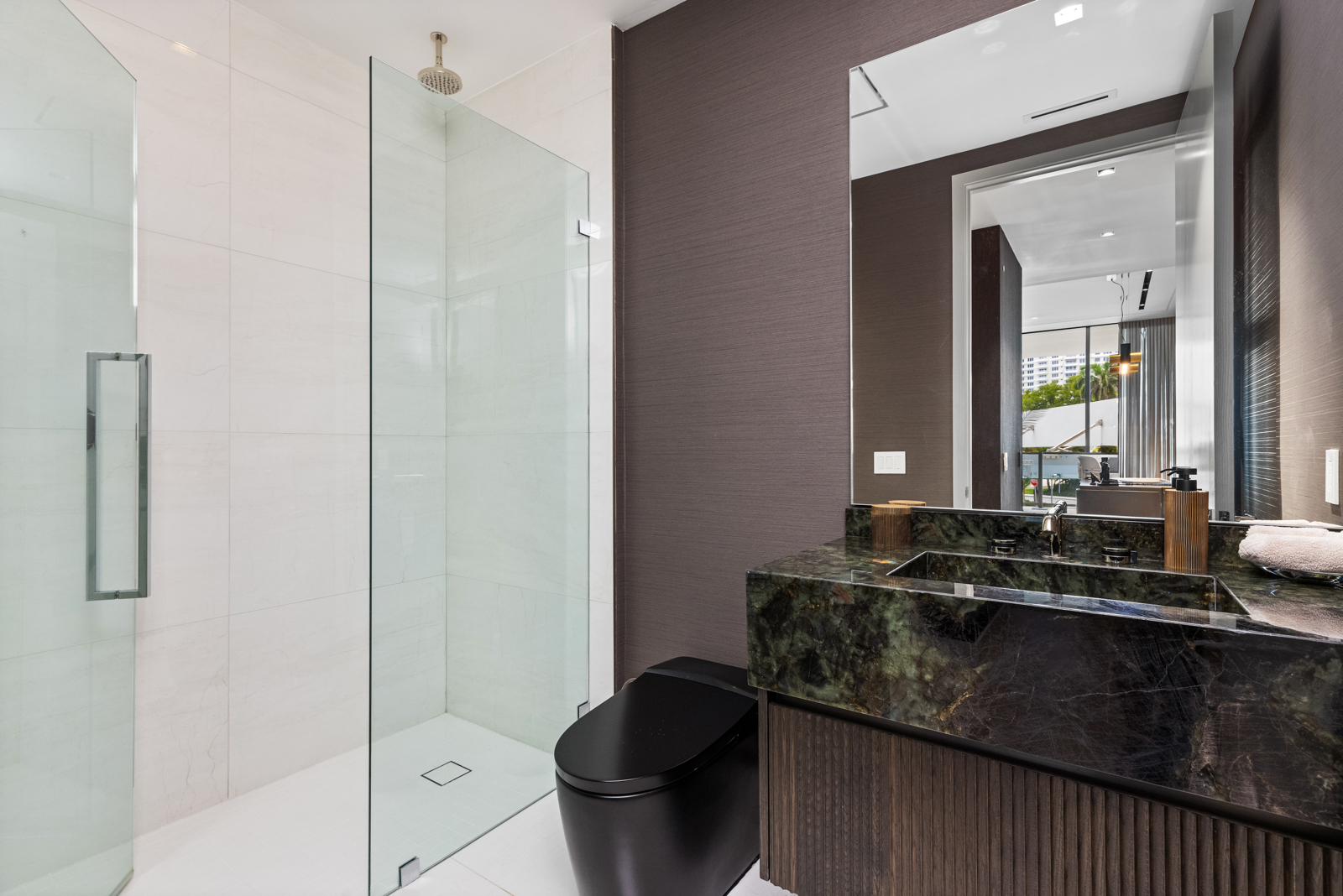This is a modern primary bathroom featuring a glass-enclosed shower with white tile walls and a rainfall showerhead. The vanity has a dark green marble countertop with a dark wood base, complemented by a large mirror and dark brown textured wallpaper. A sleek black toilet adds a contemporary touch to the space.